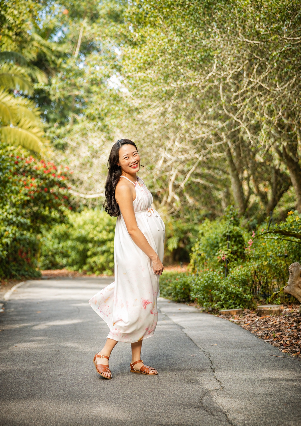 Smiling pregnant Asian woman in white floral dress posing on a lush green tree-lined path.