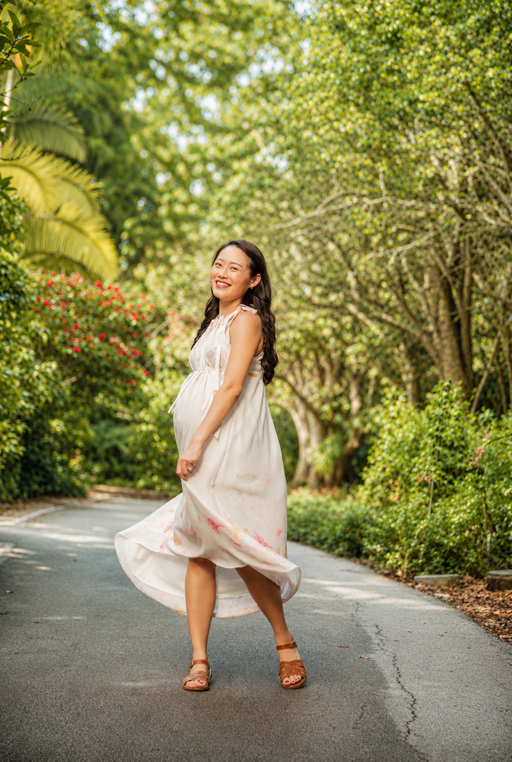 Joyful pregnant Asian woman twirling in a white dress on a sunlit tree-lined garden path.