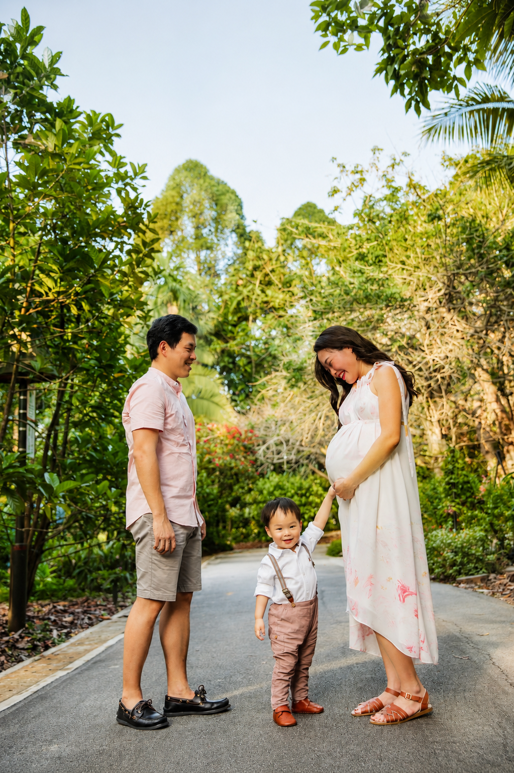 Asian family of three sharing a tender moment on a sunlit garden path during maternity shoot.