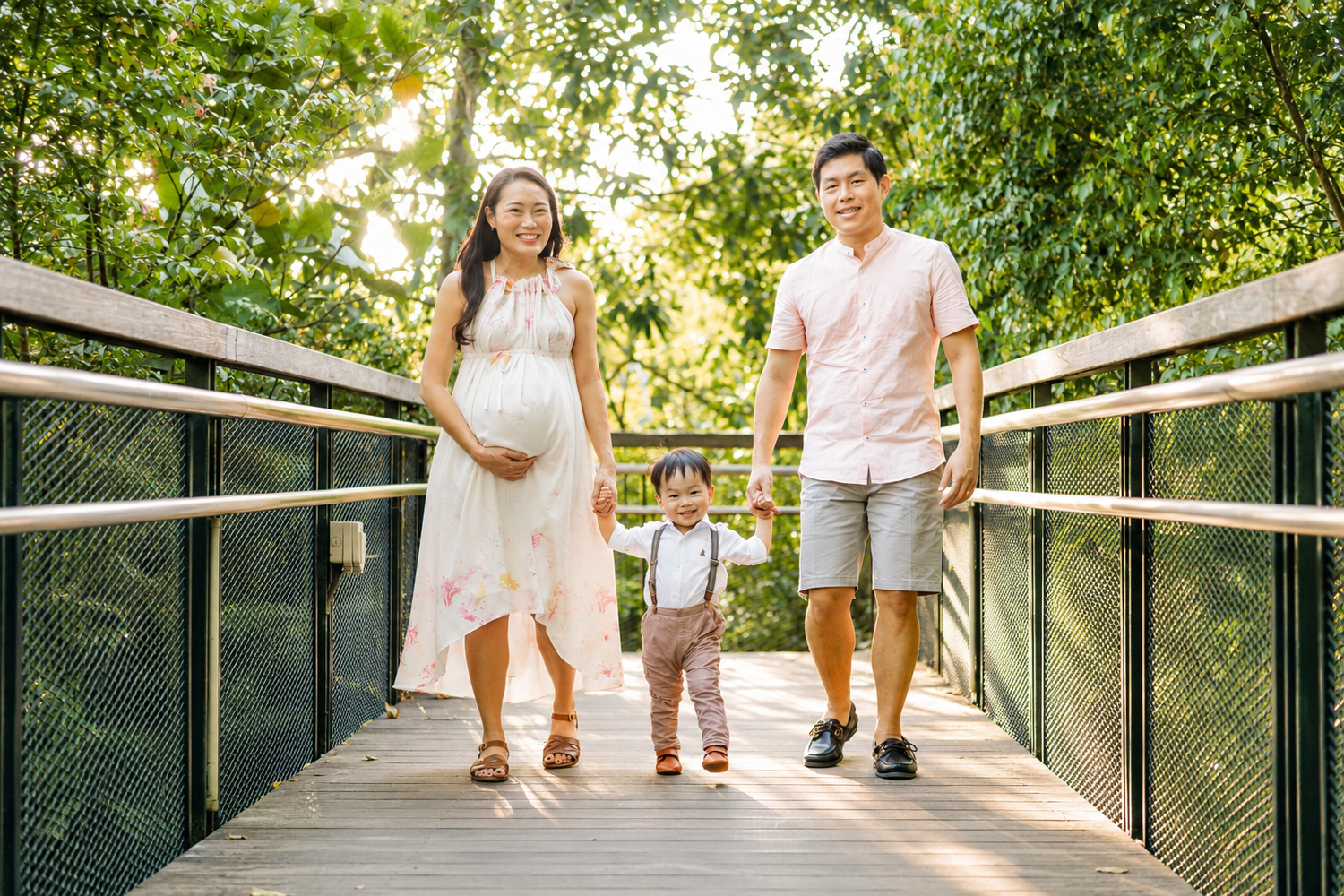 Happy Asian family with pregnant mother, father, and toddler walking on a wooden bridge.