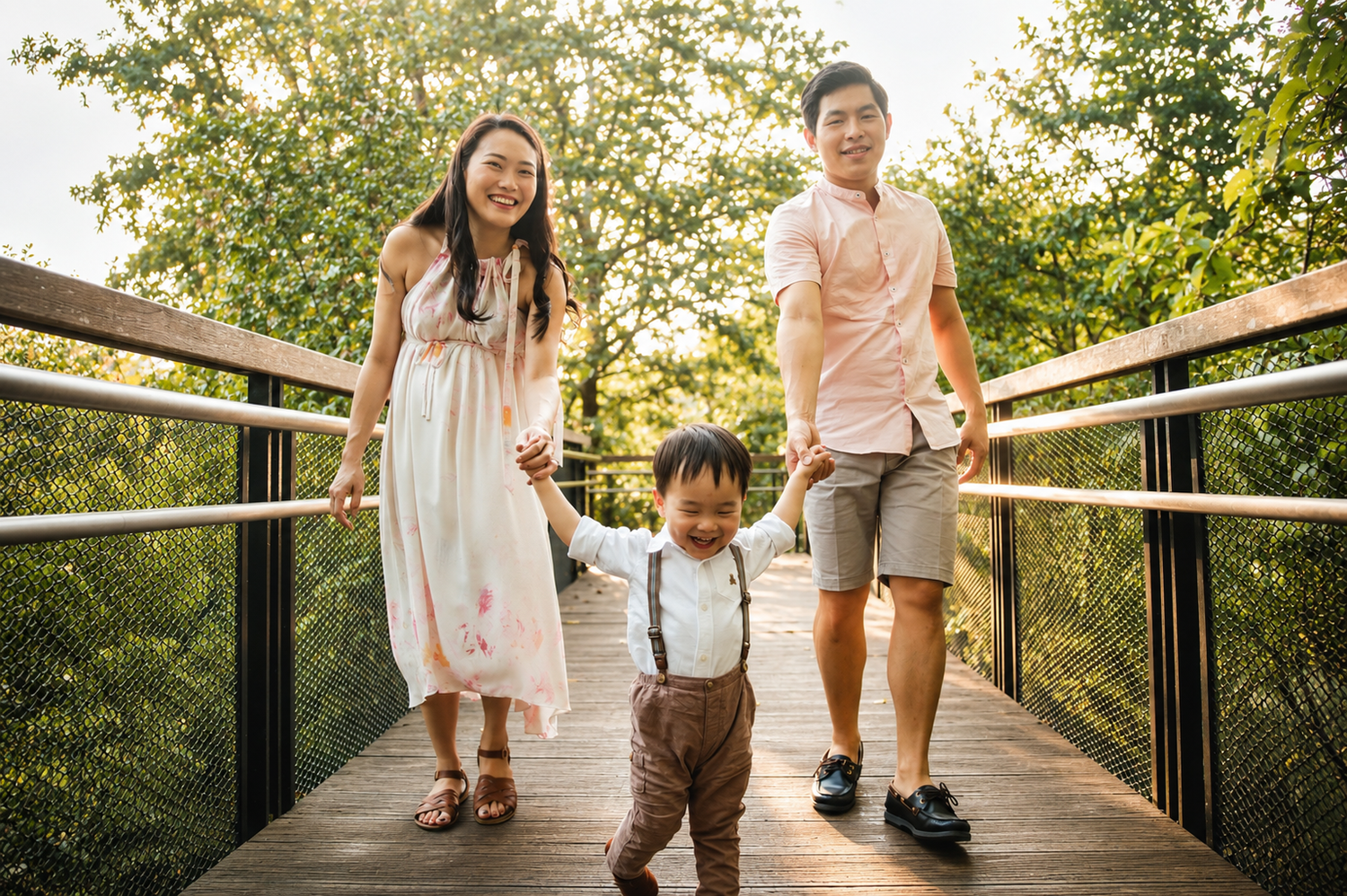 Smiling Asian family holding toddler son's hands while walking on a sunlit wooden bridge.