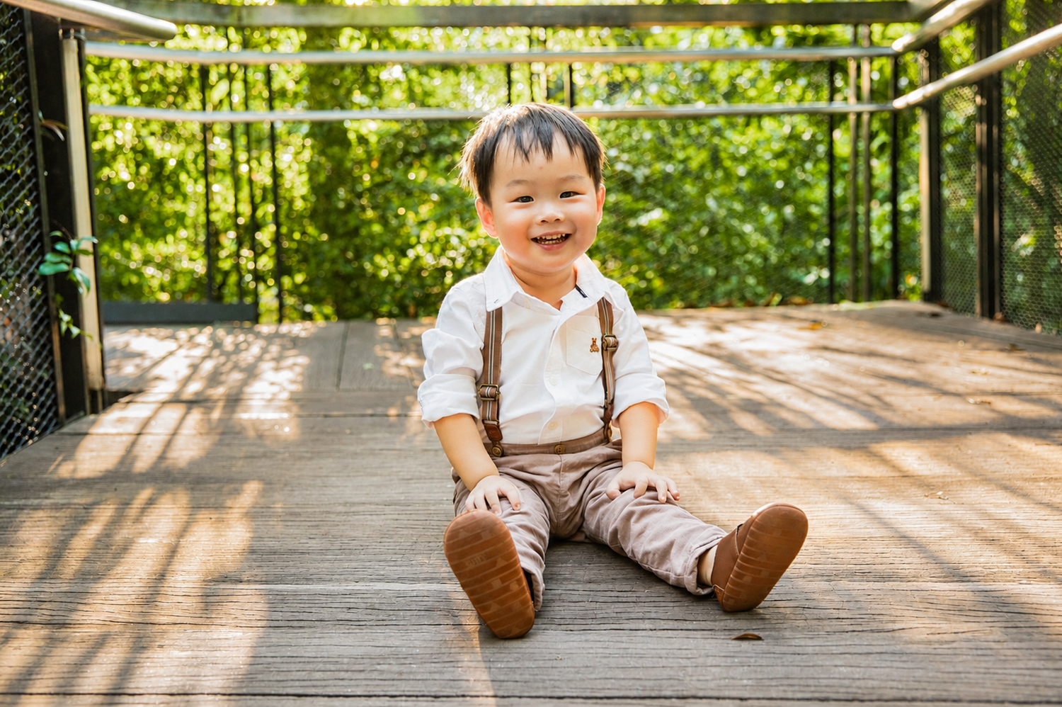 Adorable smiling toddler boy in suspenders sitting on a sunny wooden bridge surrounded by trees.