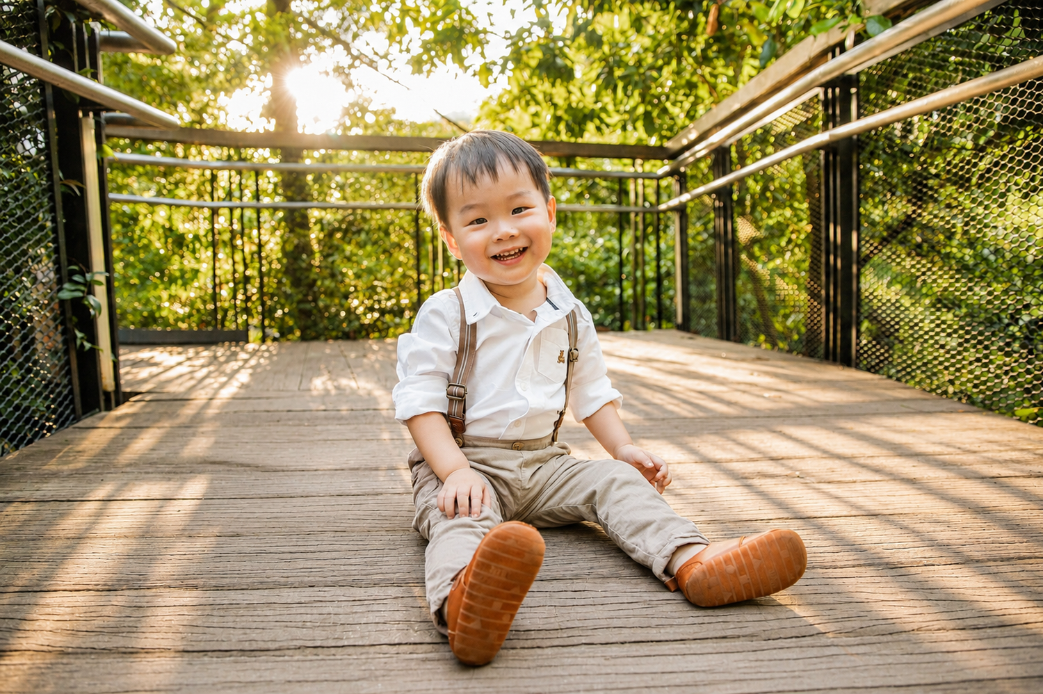 Happy toddler boy in white shirt and suspenders sitting on a wooden bridge in golden sunlight.