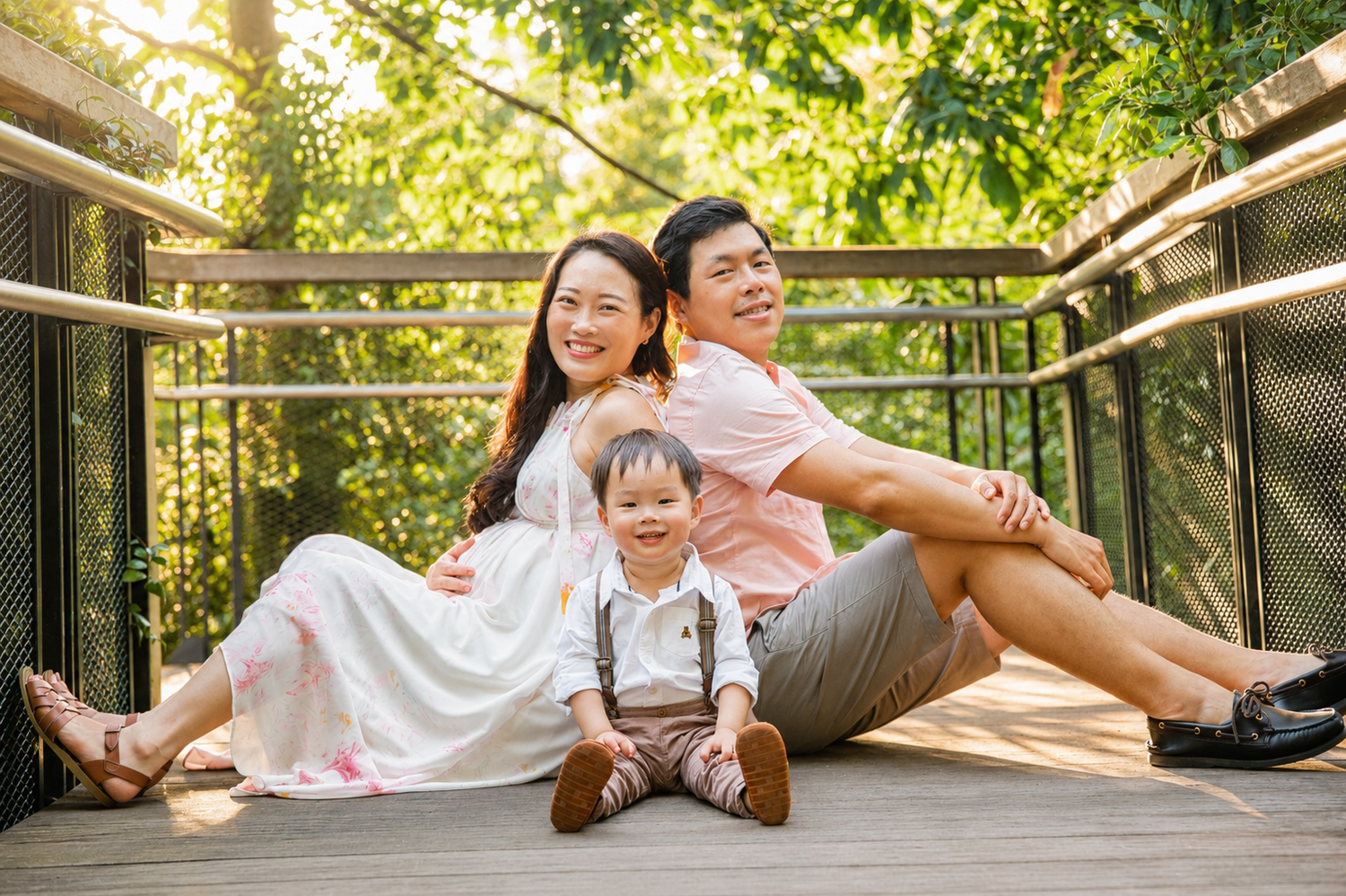 Asian family of three sitting together on a wooden bridge surrounded by lush green trees.