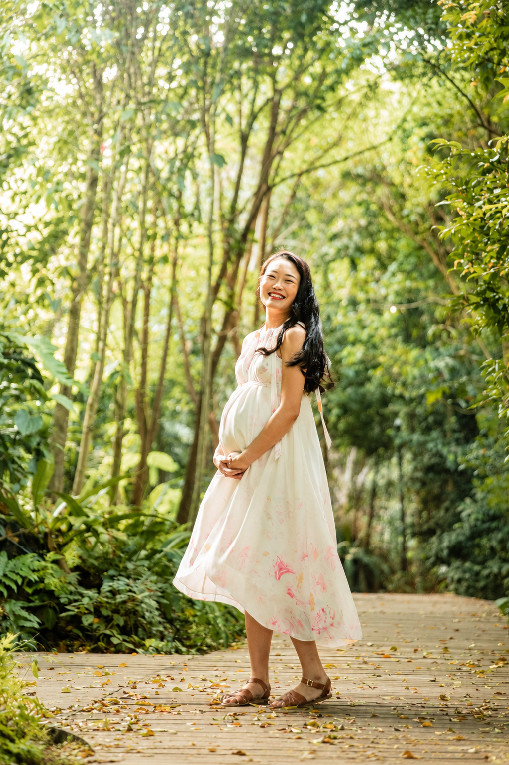 Laughing pregnant Asian woman in white floral dress standing on a sunlit wooden forest path.