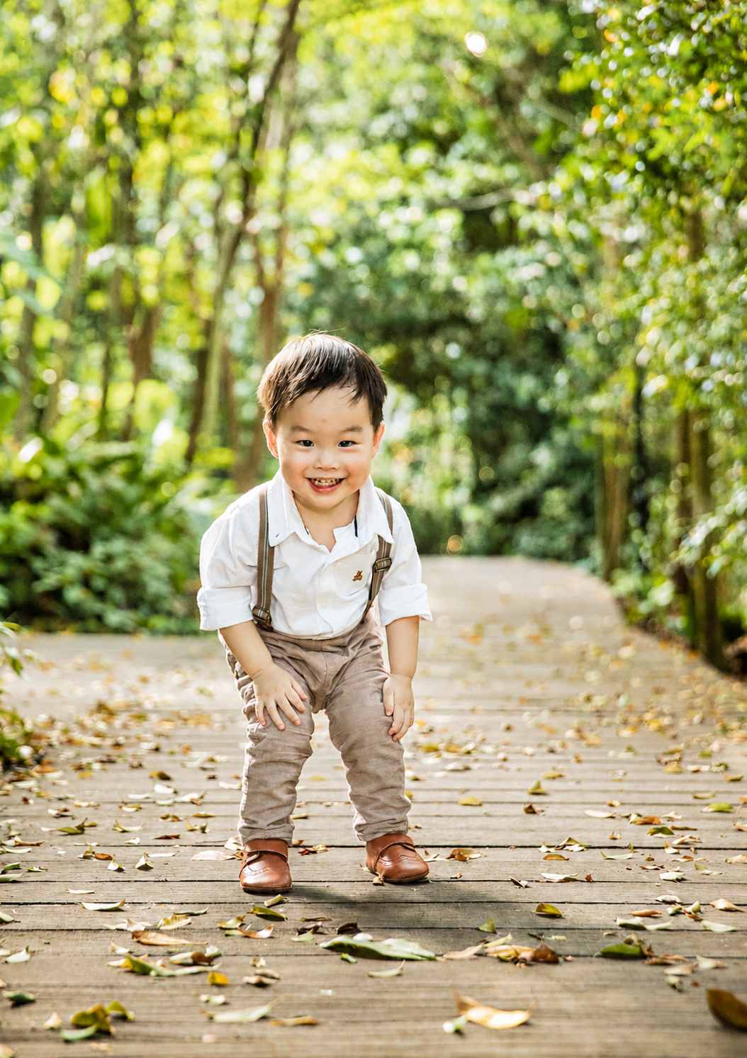 Happy Asian toddler boy in suspenders smiles on a wooden path surrounded by lush greenery.