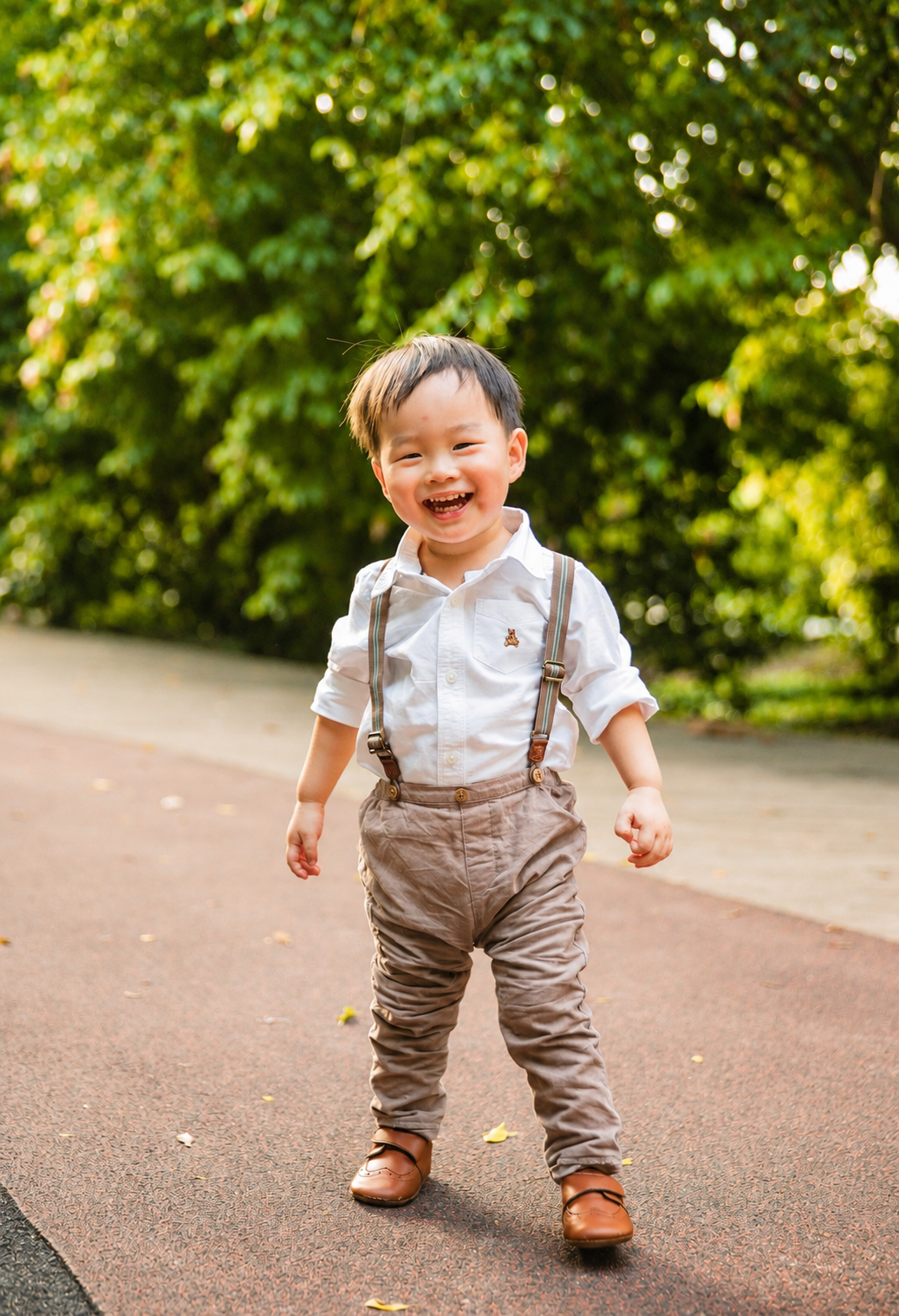 Cheerful Asian toddler boy in white shirt and suspenders laughs while standing on a sunny path.