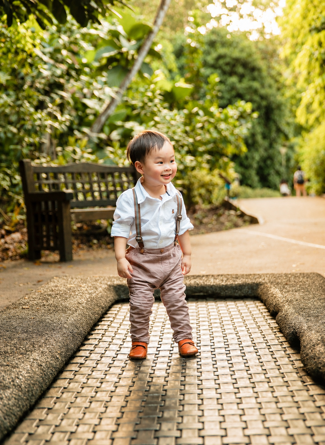 Adorable Asian toddler in suspenders stands on a garden path smiling with green trees behind him.