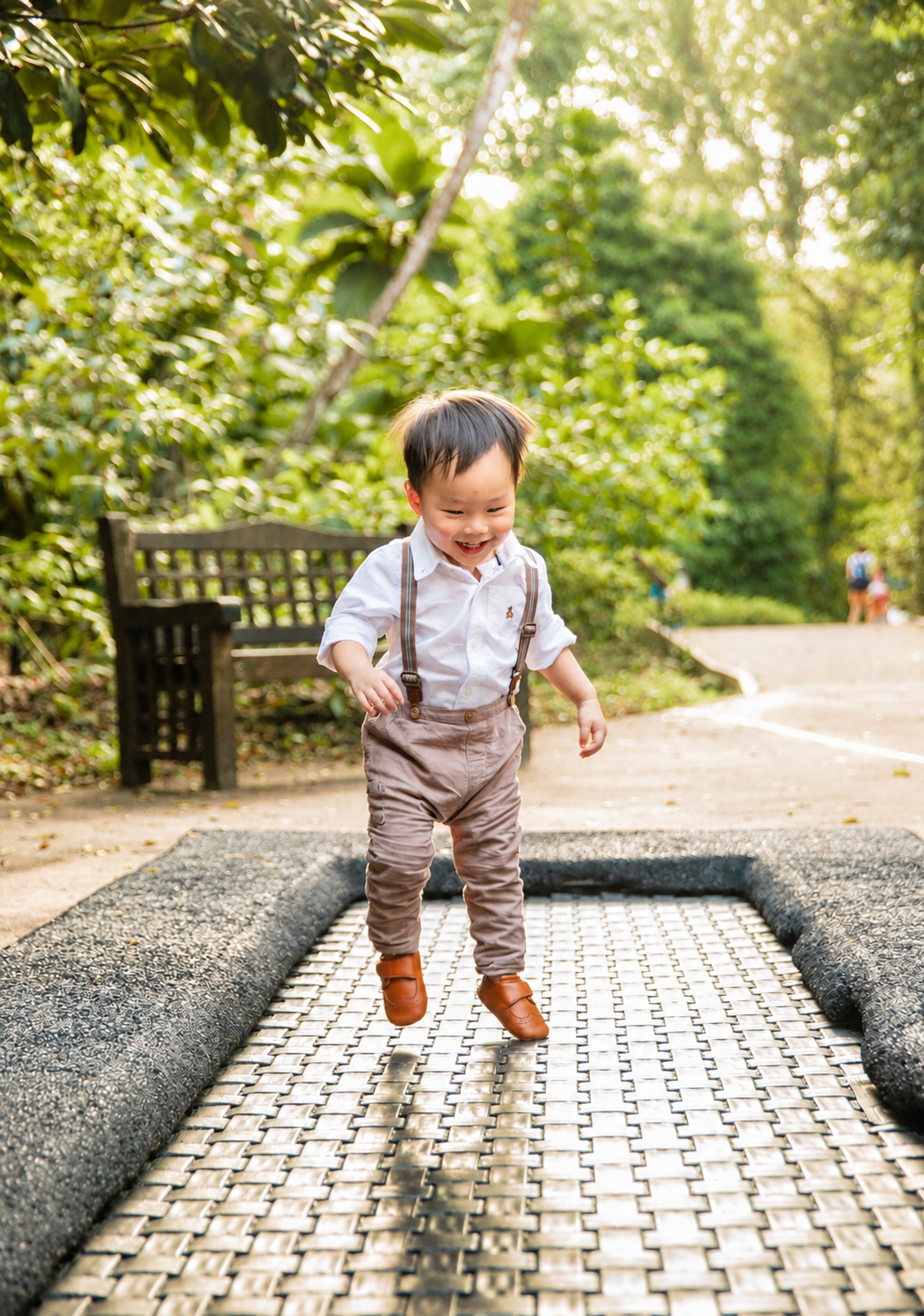 Joyful Asian toddler boy in suspenders jumps on a garden path with lush green trees in background.
