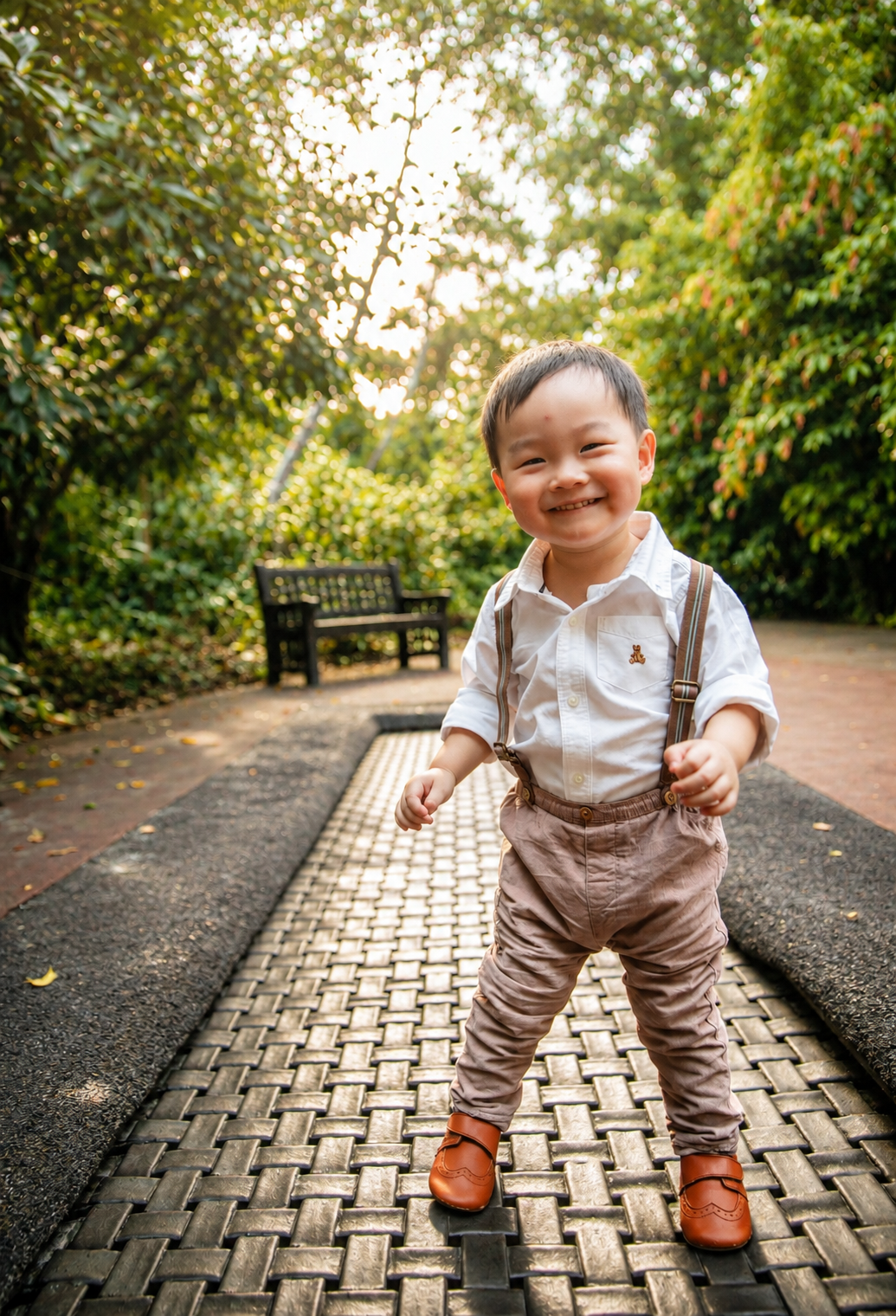 Smiling Asian toddler boy in white shirt and suspenders poses playfully on a sunlit garden path.