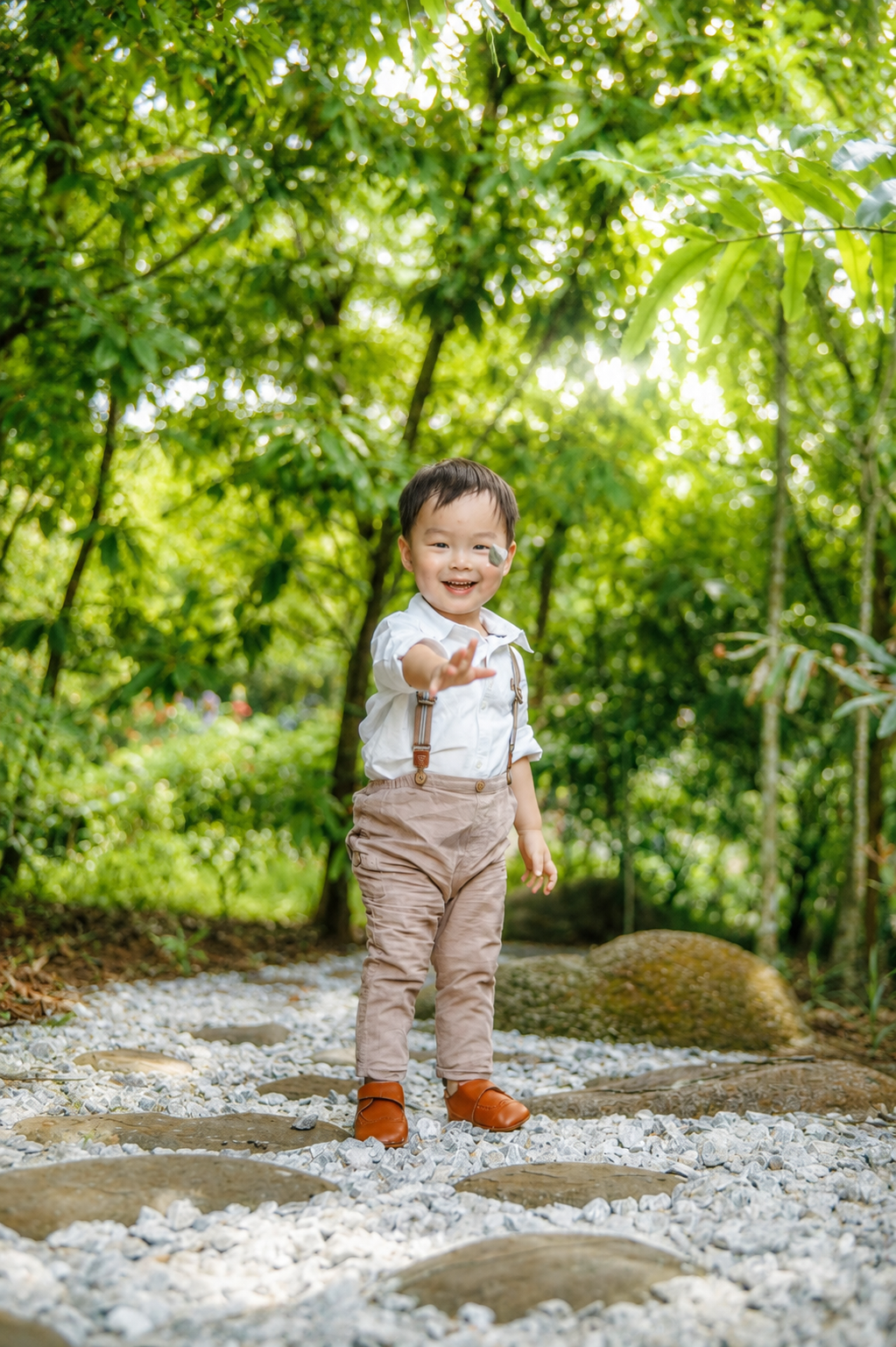 Happy Asian toddler boy in suspenders reaches out on a scenic stone garden path with green trees.