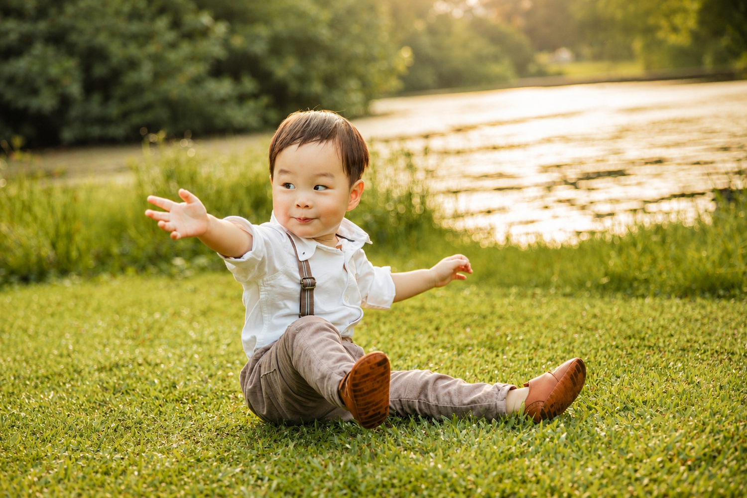 Cute Asian toddler boy in suspenders sits on green grass with arms outstretched in golden sunlight.