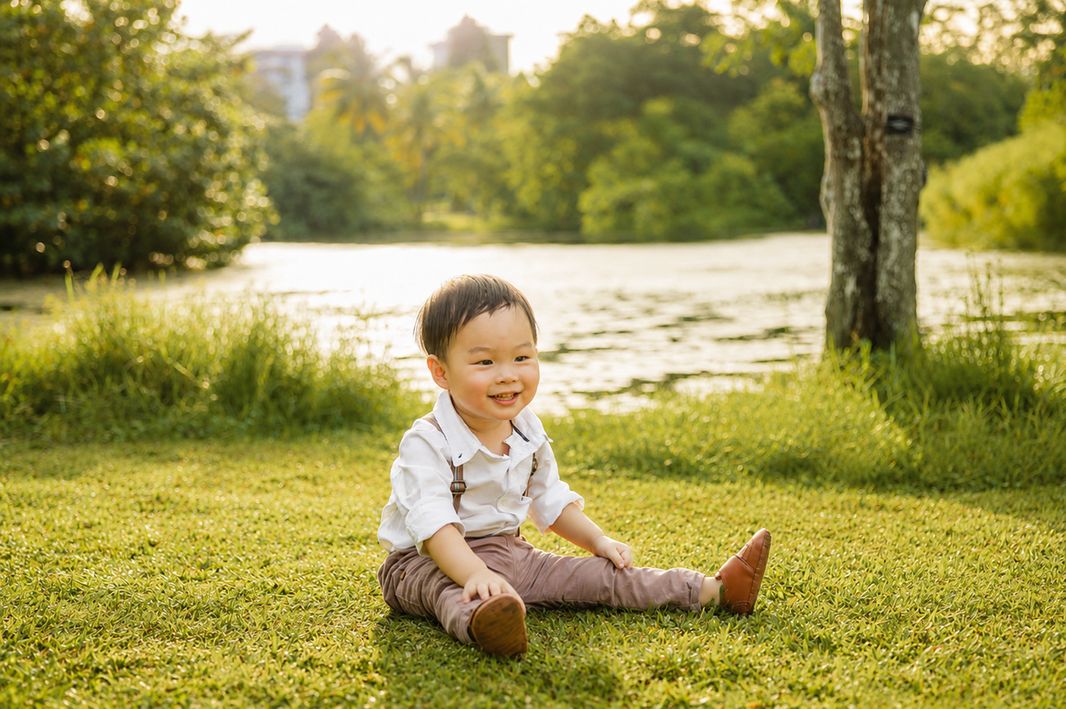 Sweet Asian toddler boy in suspenders sits smiling on lush green grass near a sunlit pond.