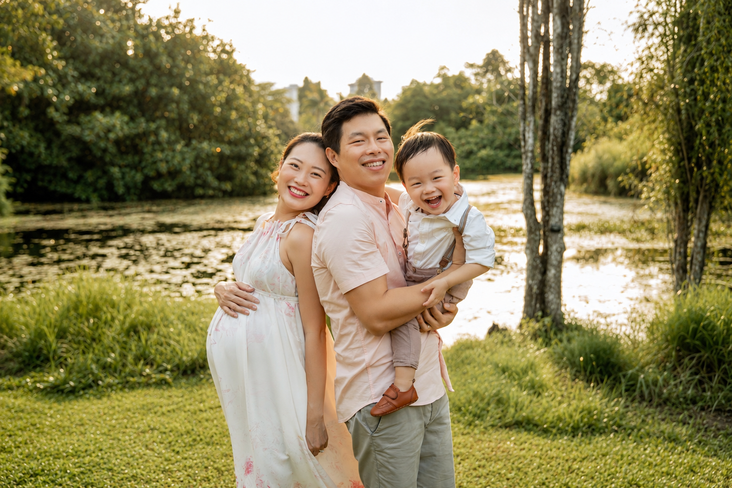 Happy Asian family with pregnant mother, father, and toddler son posing outdoors by a pond.