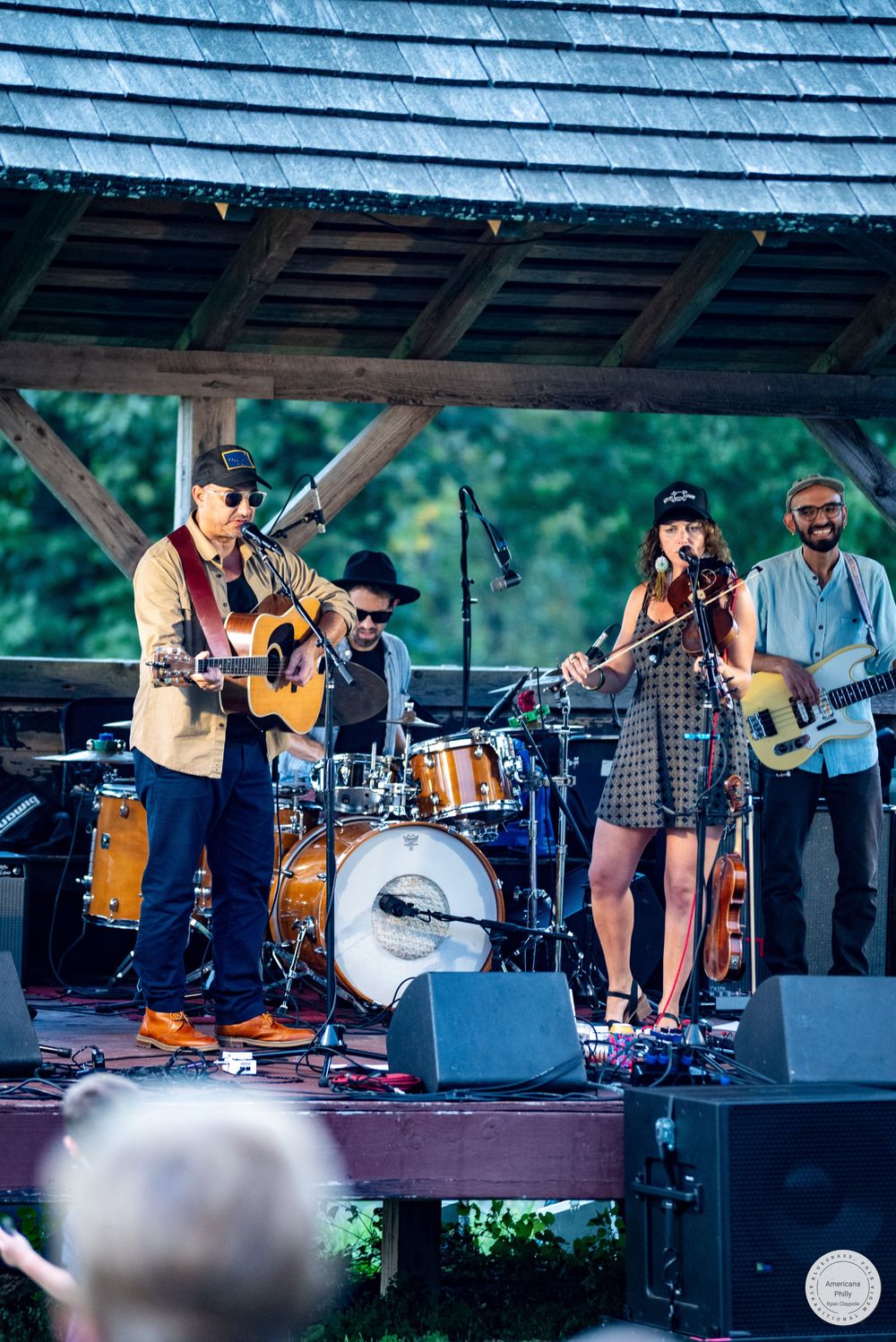 Live band performing on outdoor stage featuring guitarist, drummer, female vocalist, and bassist at a music festival.