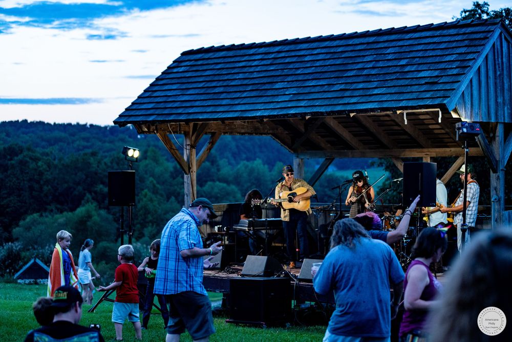 Outdoor folk band performs on rustic wooden stage at dusk with crowd gathered on green lawn at a summer festival.