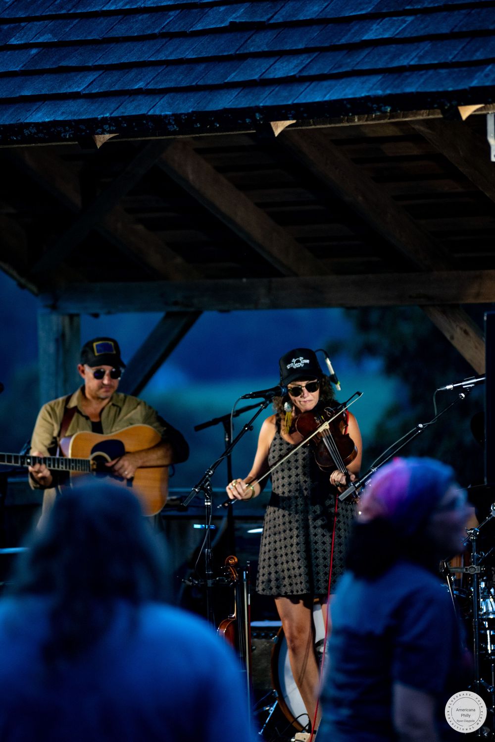 Woman playing violin and man playing acoustic guitar perform outdoors on a rustic wooden stage at a live music event.