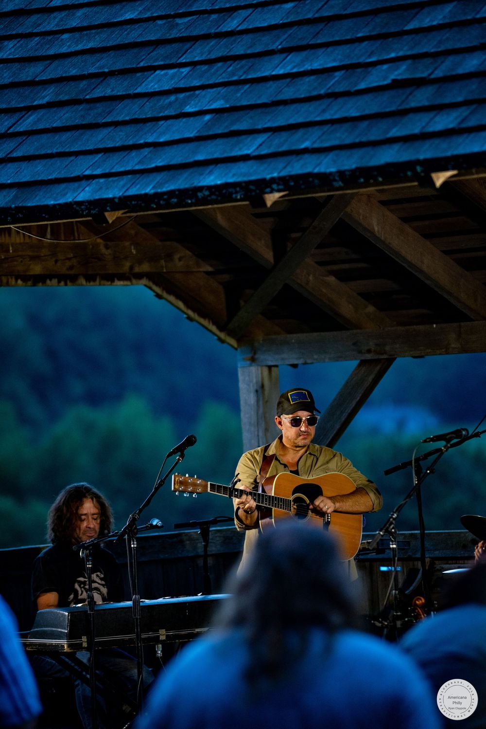 Musician plays acoustic guitar on outdoor stage under wooden roof with green forested hills at dusk.