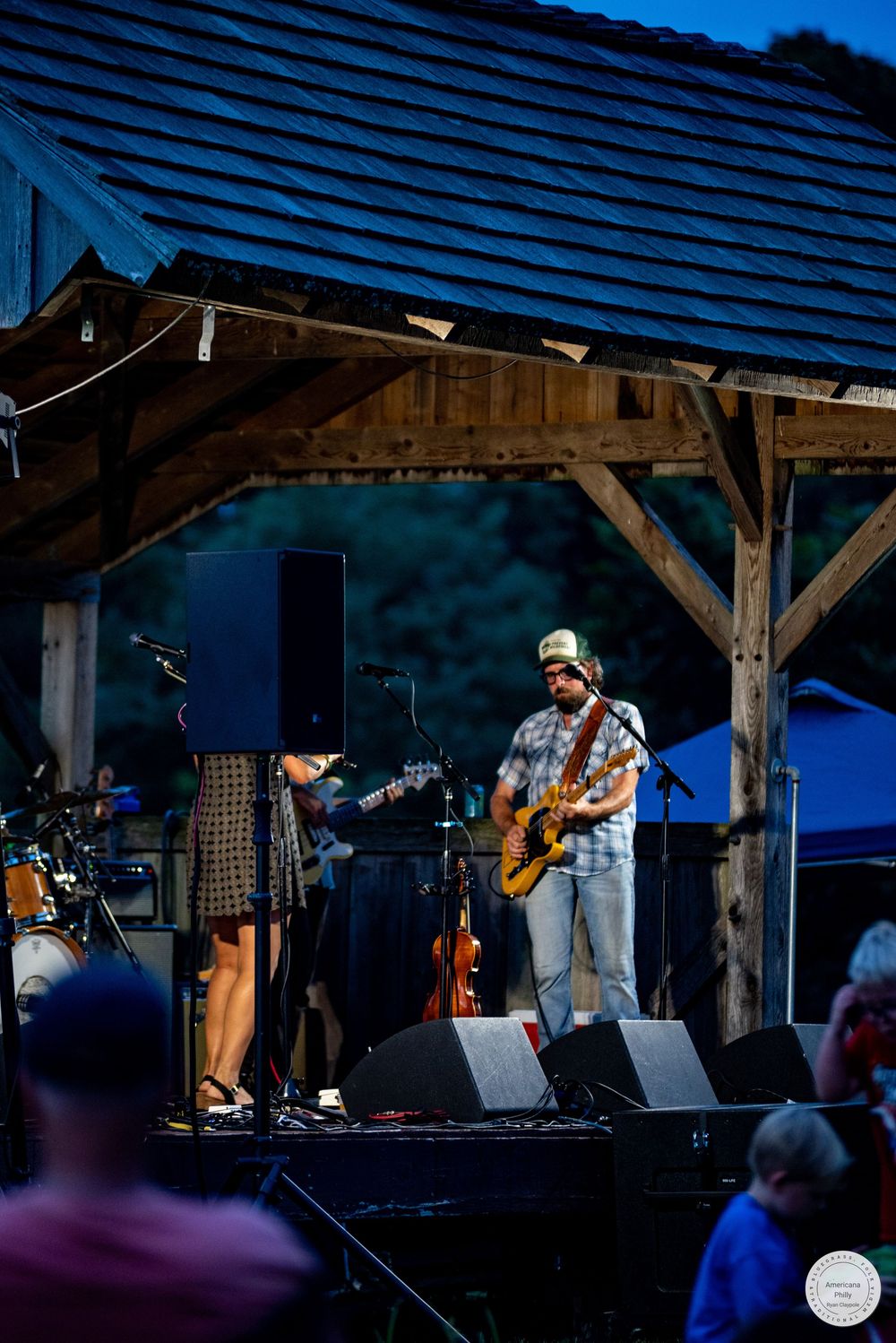 Outdoor evening concert with two musicians performing on a rustic wooden stage, one playing guitar and one singing.