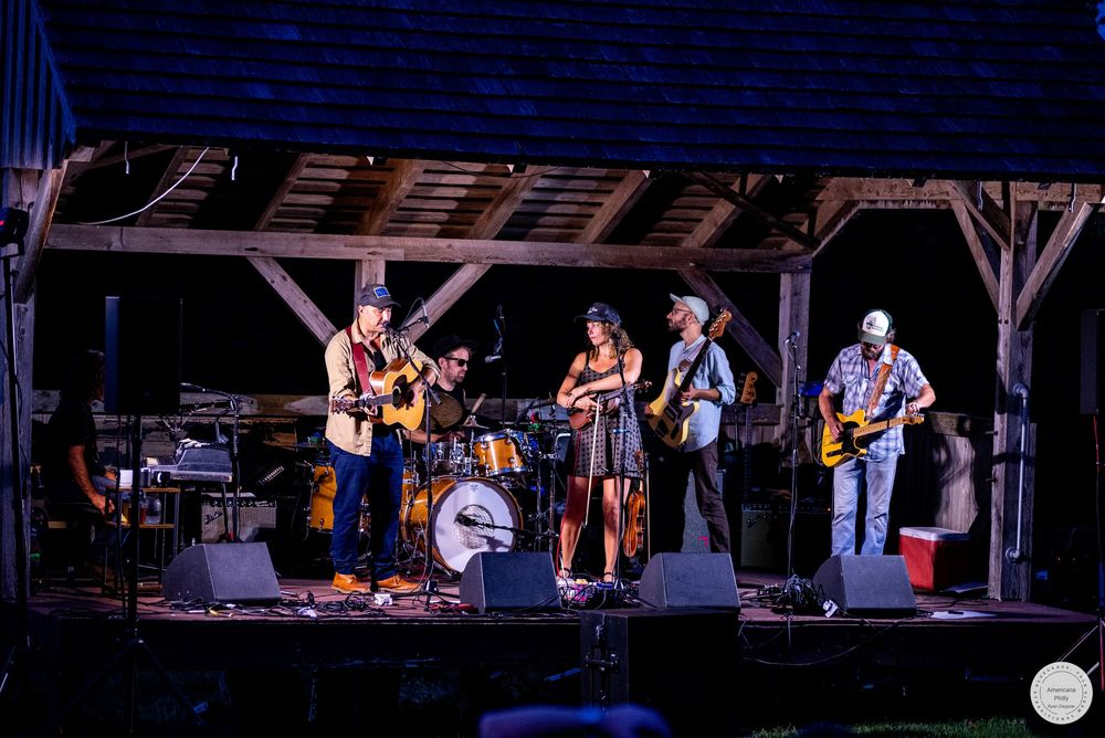 A live country band performs on an outdoor rustic wooden stage at night, with musicians playing guitar and drums.