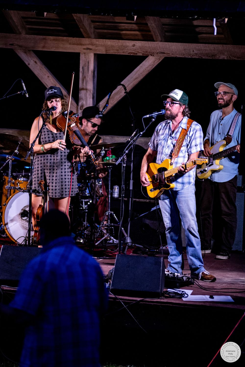 Live band performing on rustic wooden stage featuring violin, electric guitars, and drums at outdoor music festival.