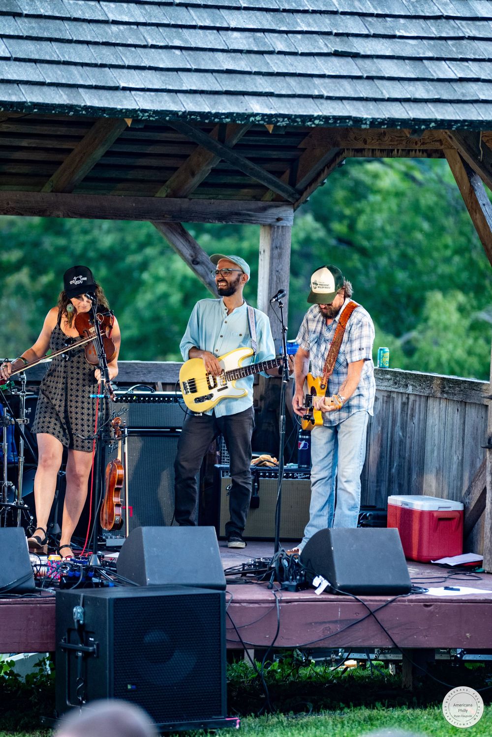 Three musicians perform on an outdoor stage under a wooden pavilion, with two guitarists and a female vocalist at a live music event.