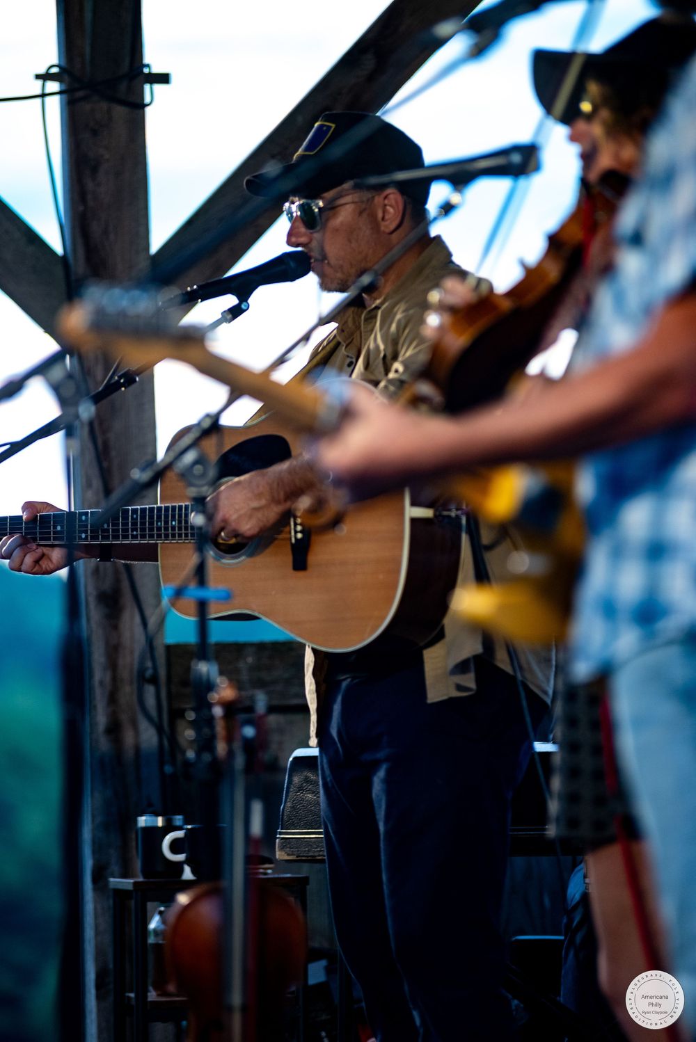 Man in cowboy hat playing acoustic guitar on stage at outdoor bluegrass music festival with band members behind him.