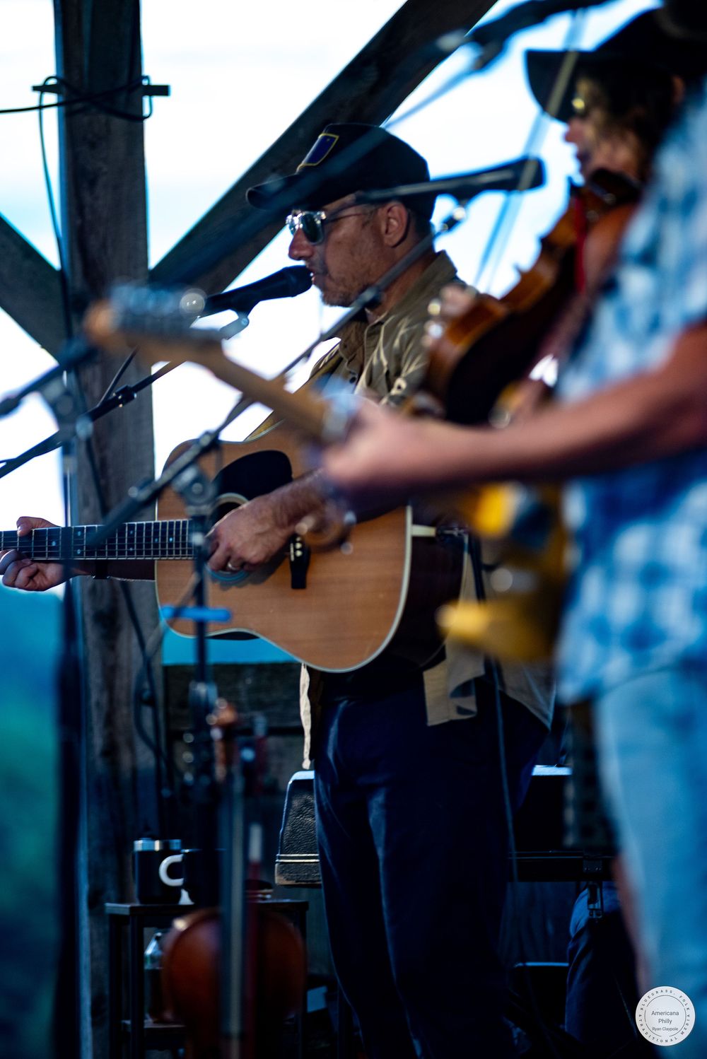 Musician playing acoustic guitar on stage wearing a black cowboy hat and sunglasses at an outdoor concert.