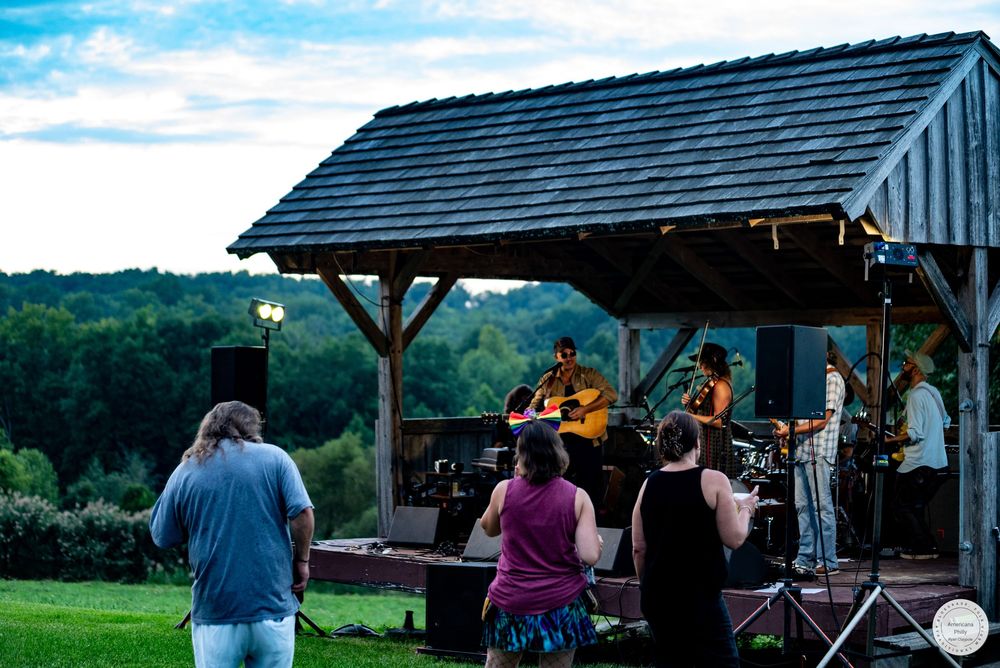Outdoor live band performance under a rustic wooden pavilion at dusk, with audience members enjoying music on a green hillside.