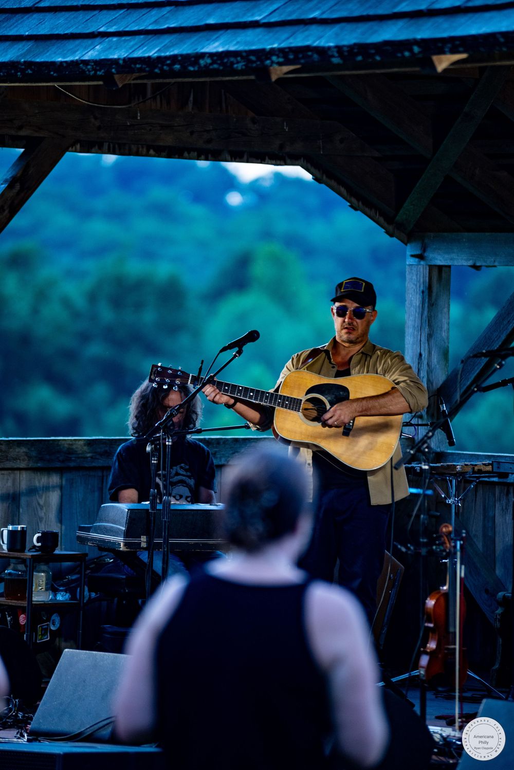 Musician playing acoustic guitar on outdoor stage under wooden pavilion with lush green trees in background.