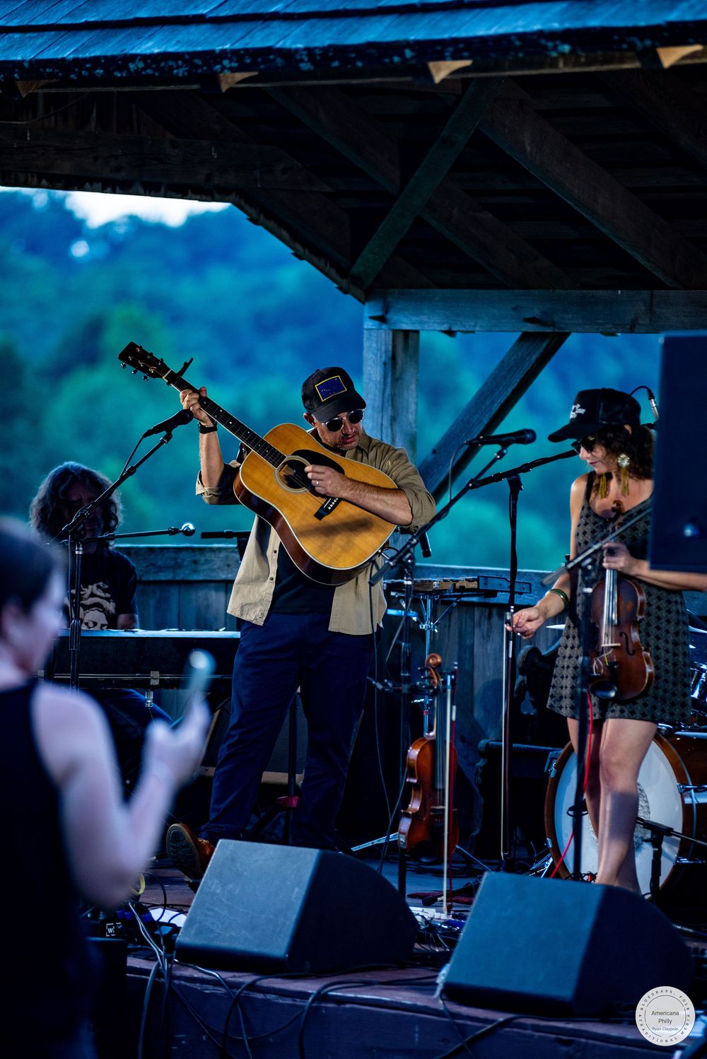 Outdoor folk band performs on rustic wooden stage with guitarist, fiddler, and vocalist against lush green backdrop.