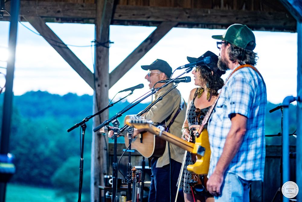 Three musicians perform on an outdoor rustic stage at dusk, with a guitarist, female vocalist, and bassist playing country music.
