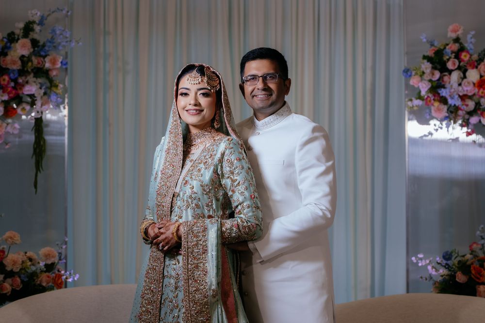 Smiling bride in an embroidered teal and gold outfit with a dupatta stands beside a groom in a white kurta, surrounded by pink floral decor.