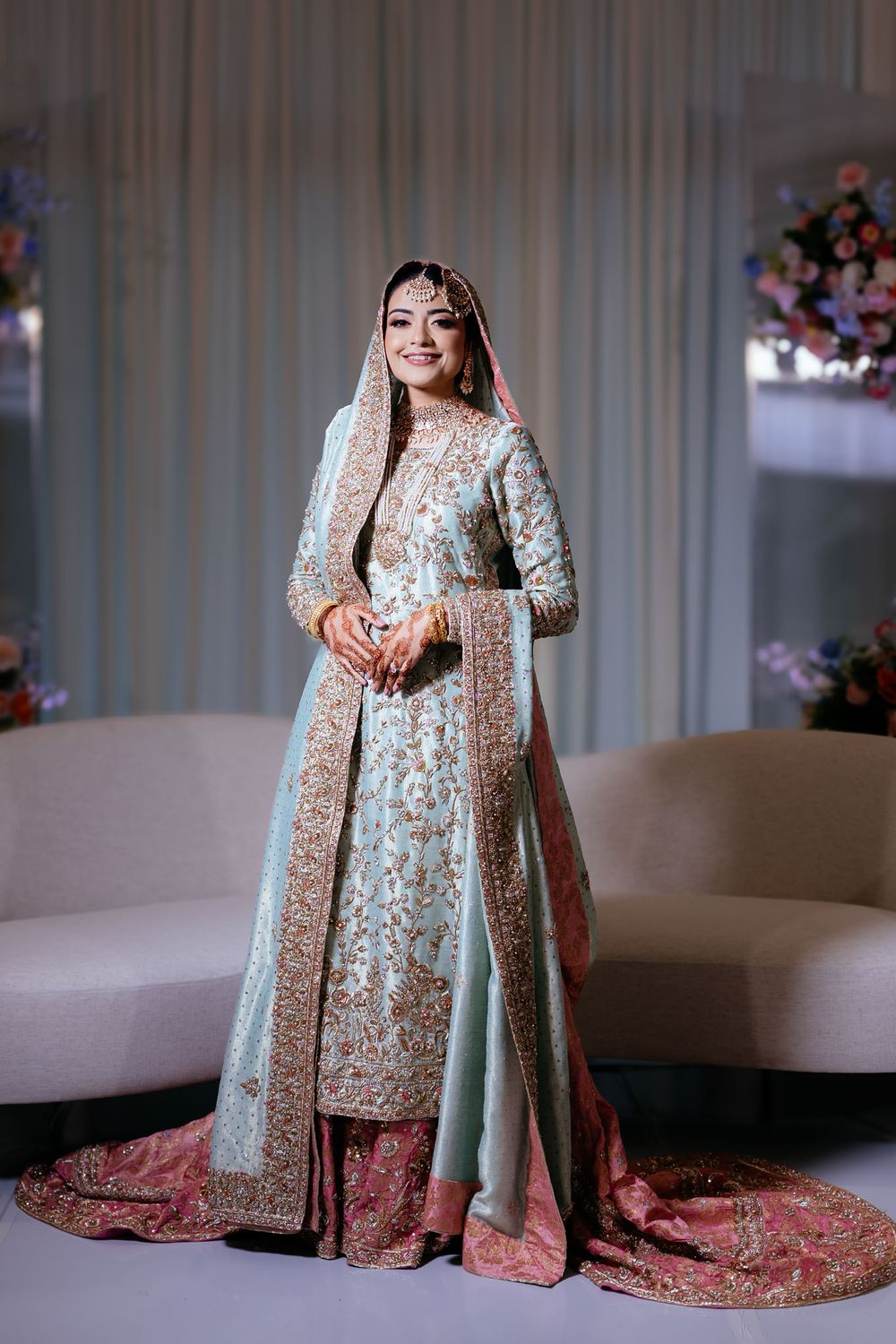 Bride in a pale blue embroidered South Asian wedding outfit with a pink gold-trimmed dupatta and long train, standing beside a cream sofa.