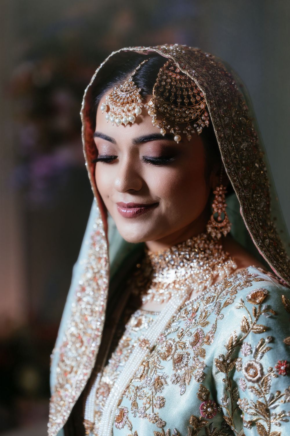 Bride in a pale blue embroidered lehenga with gold dupatta, matha patti, kundan necklace and earrings, looking down with a soft smile.