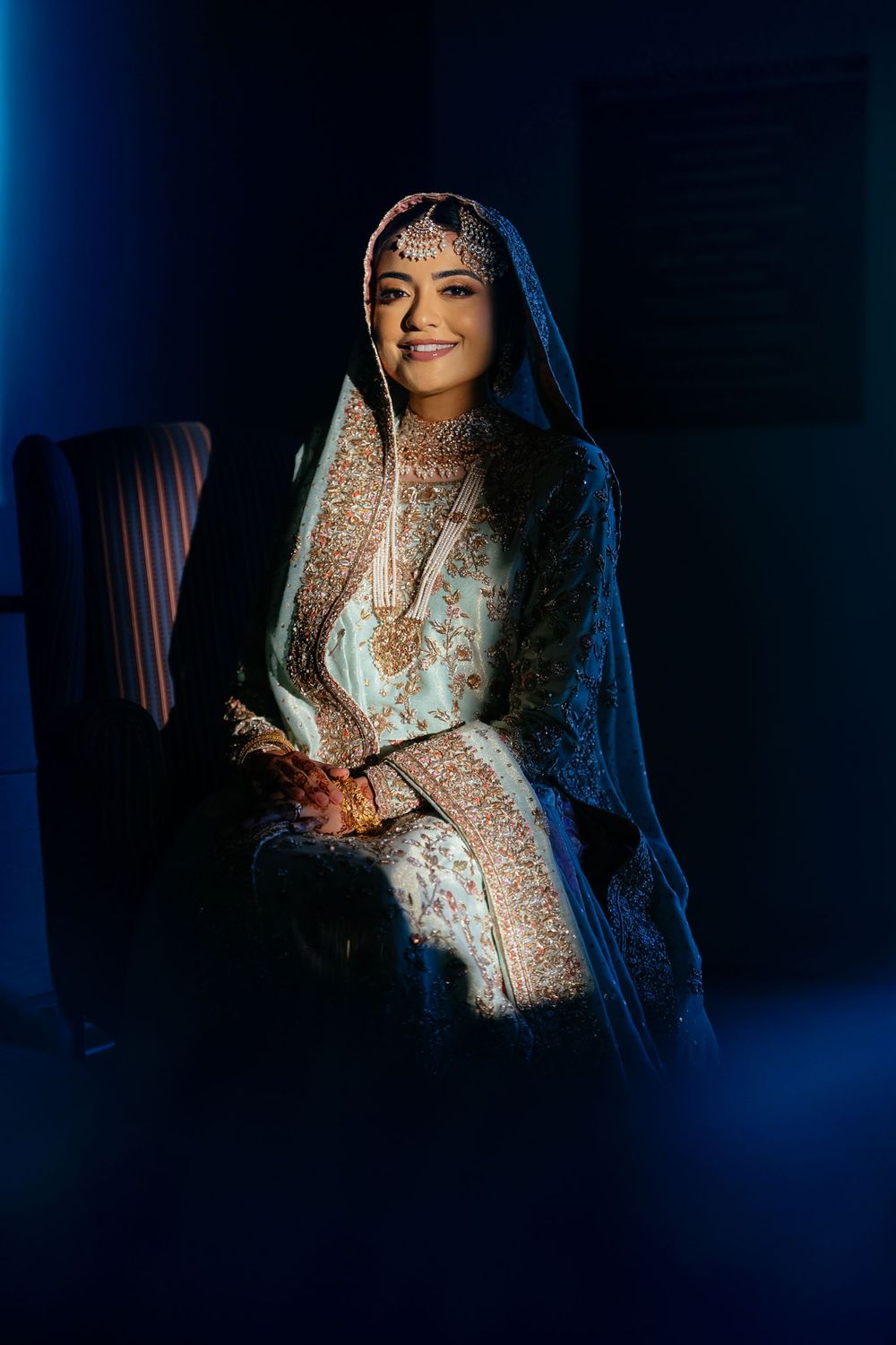 Smiling bride seated in dramatic blue light, wearing an ornate pale blue and gold embroidered South Asian wedding outfit with a matha patti.