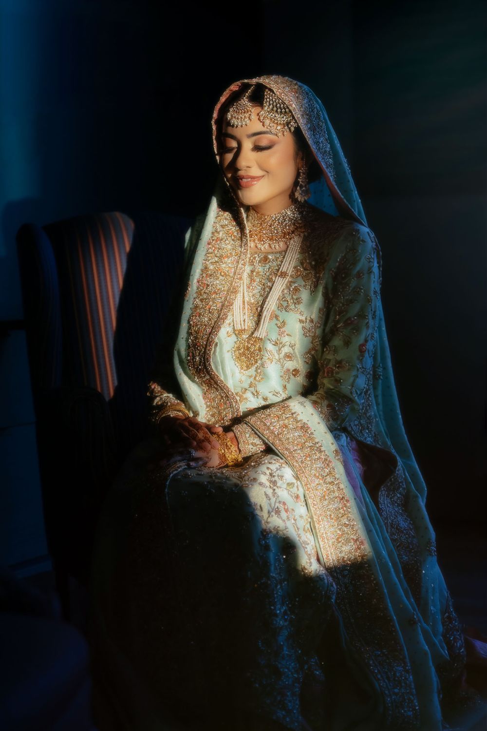Bride in an ornate teal and gold embroidered bridal outfit with a dupatta veil and jewelry sits smiling in soft dramatic window light.