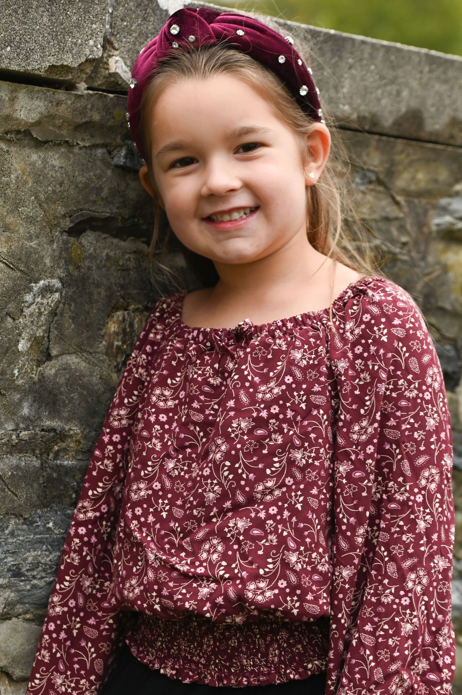 Smiling young girl in a burgundy floral peasant blouse and a matching velvet knotted headband leaning against a rustic stone wall.