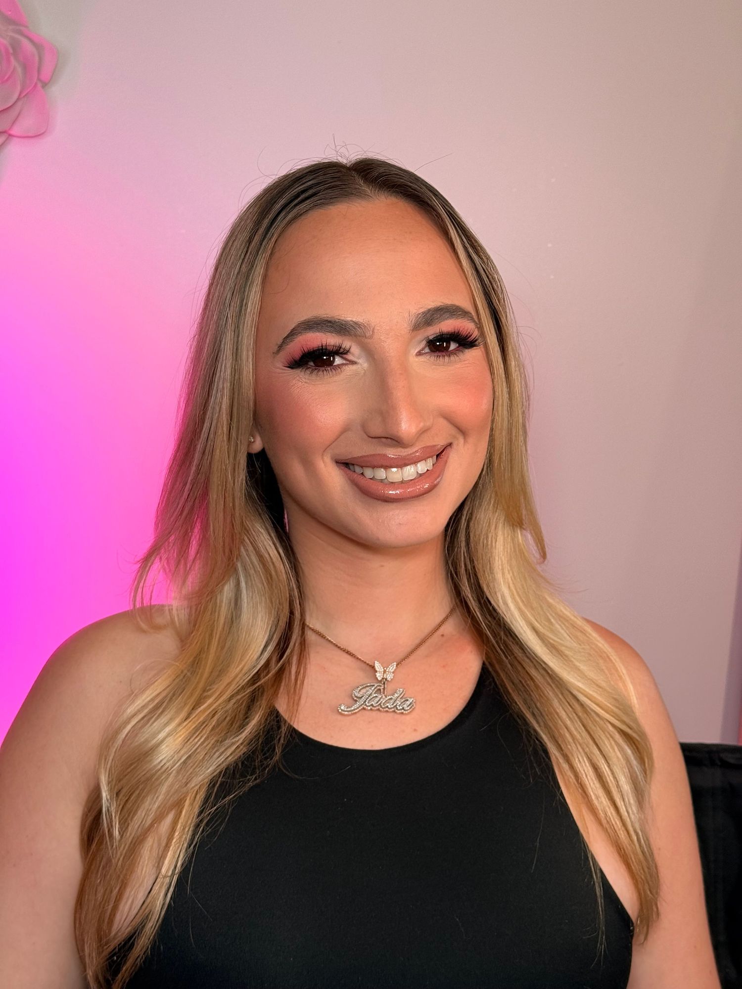 Smiling woman with long blonde hair, glam makeup and a butterfly name necklace wearing a black top against a pink-lit wall.