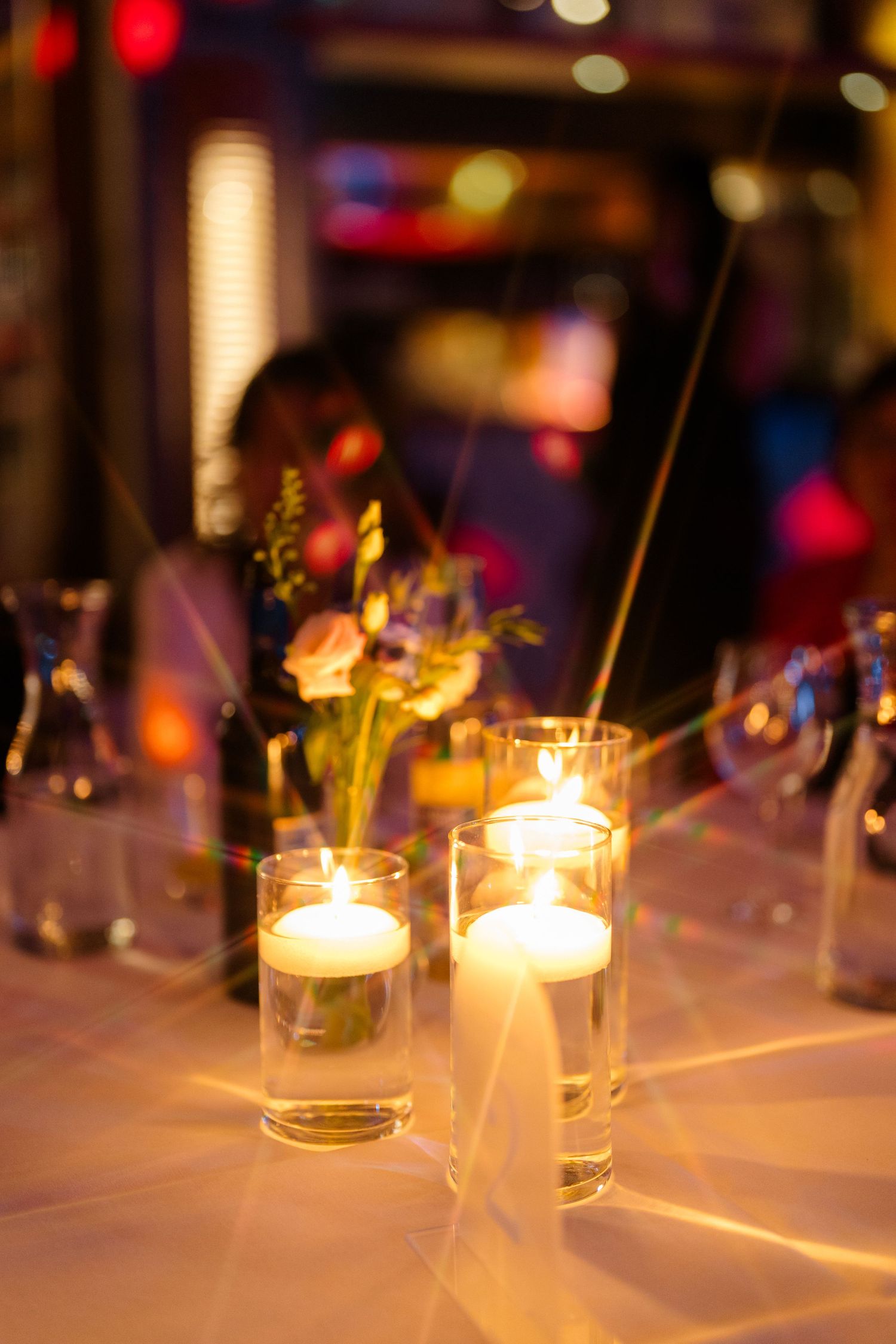 Floating candles in glass cylinders glow on a reception table beside a small floral arrangement, with warm blurred lights in the background.