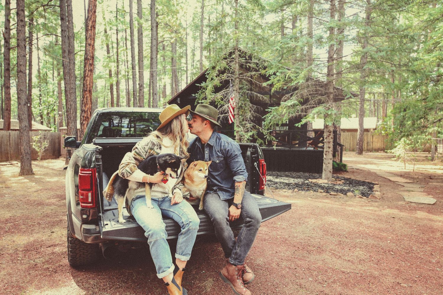 Couple in cowboy hats sitting on a pickup truck tailgate with a dog, kissing in a sunlit pine forest near a rustic log cabin.