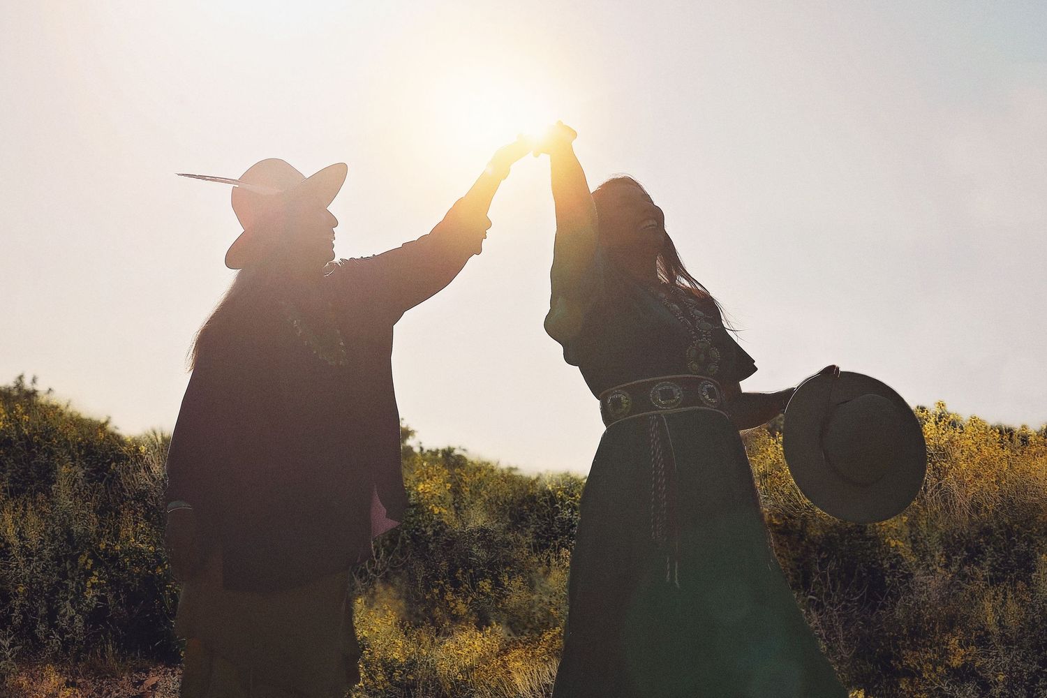 Silhouetted couple in cowboy hats hold hands raised toward the sun while dancing on a grassy hill at sunset.