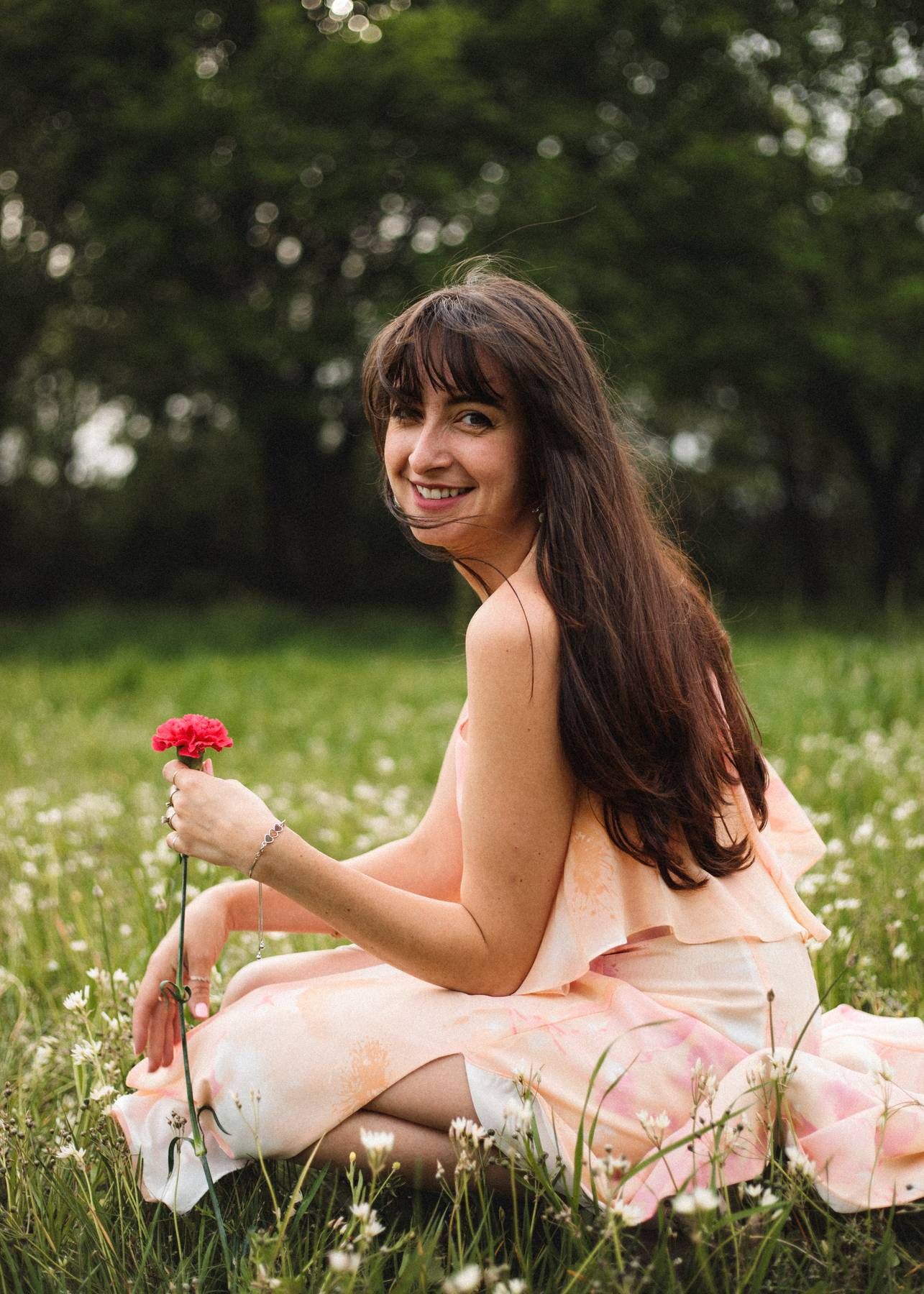 A smiling woman in a peach dress kneels in a green meadow holding a red carnation, with trees softly blurred behind her.