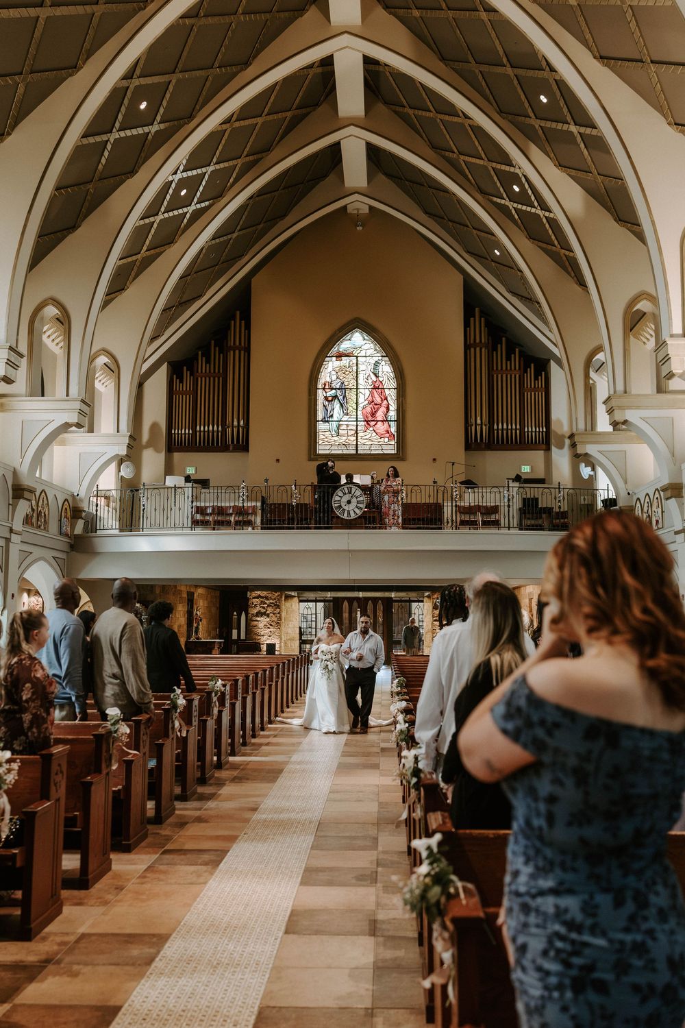 A bride and groom walk down the aisle of a grand church with arched ceilings and stained glass as guests look on.