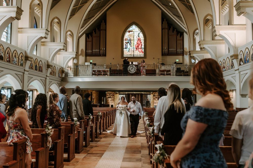 Bride and groom walk down the church aisle past seated wedding guests toward the altar and stained glass window.