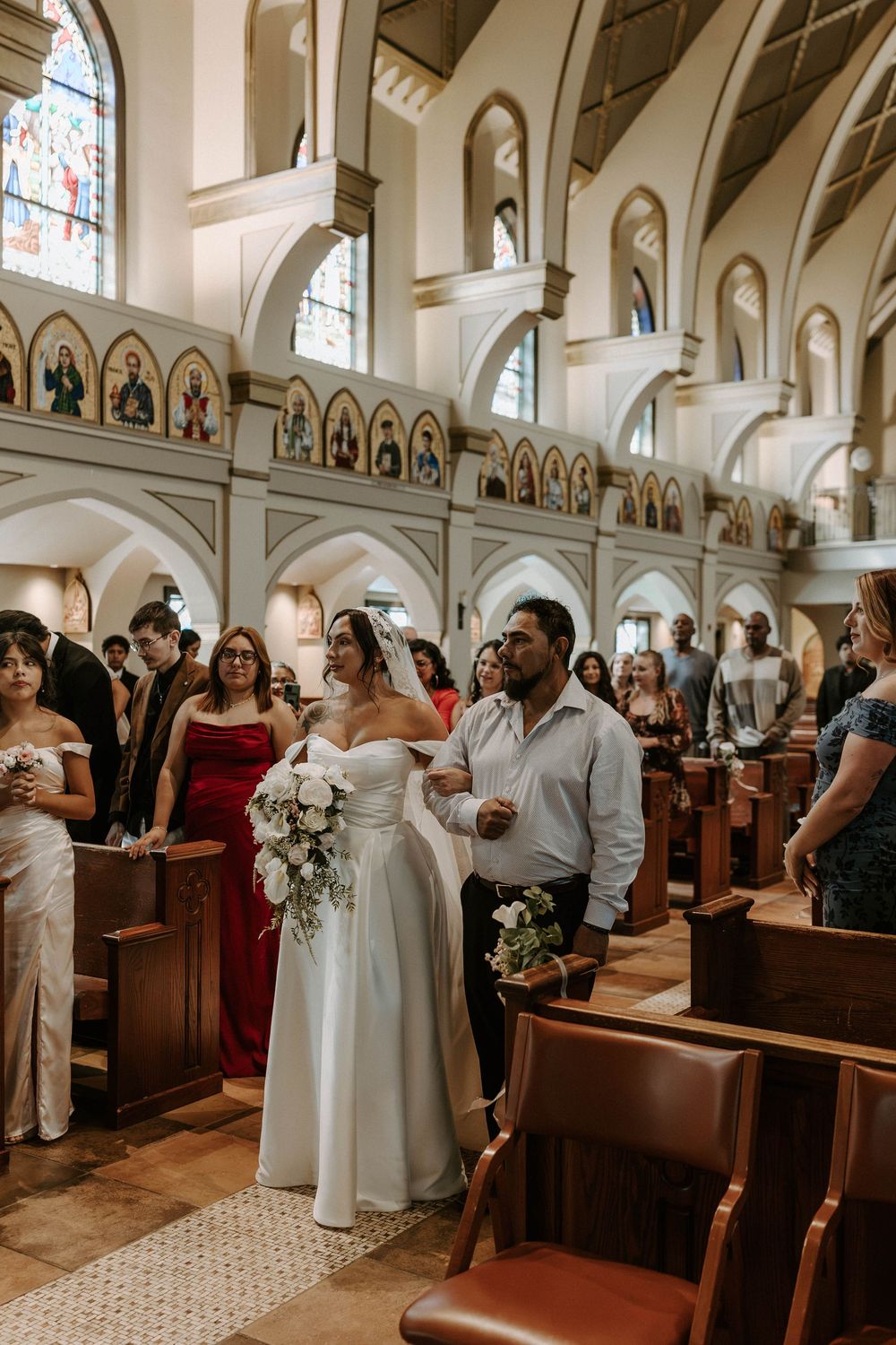 A bride in an off-the-shoulder white gown holds a bouquet while walking down the church aisle as wedding guests look on.