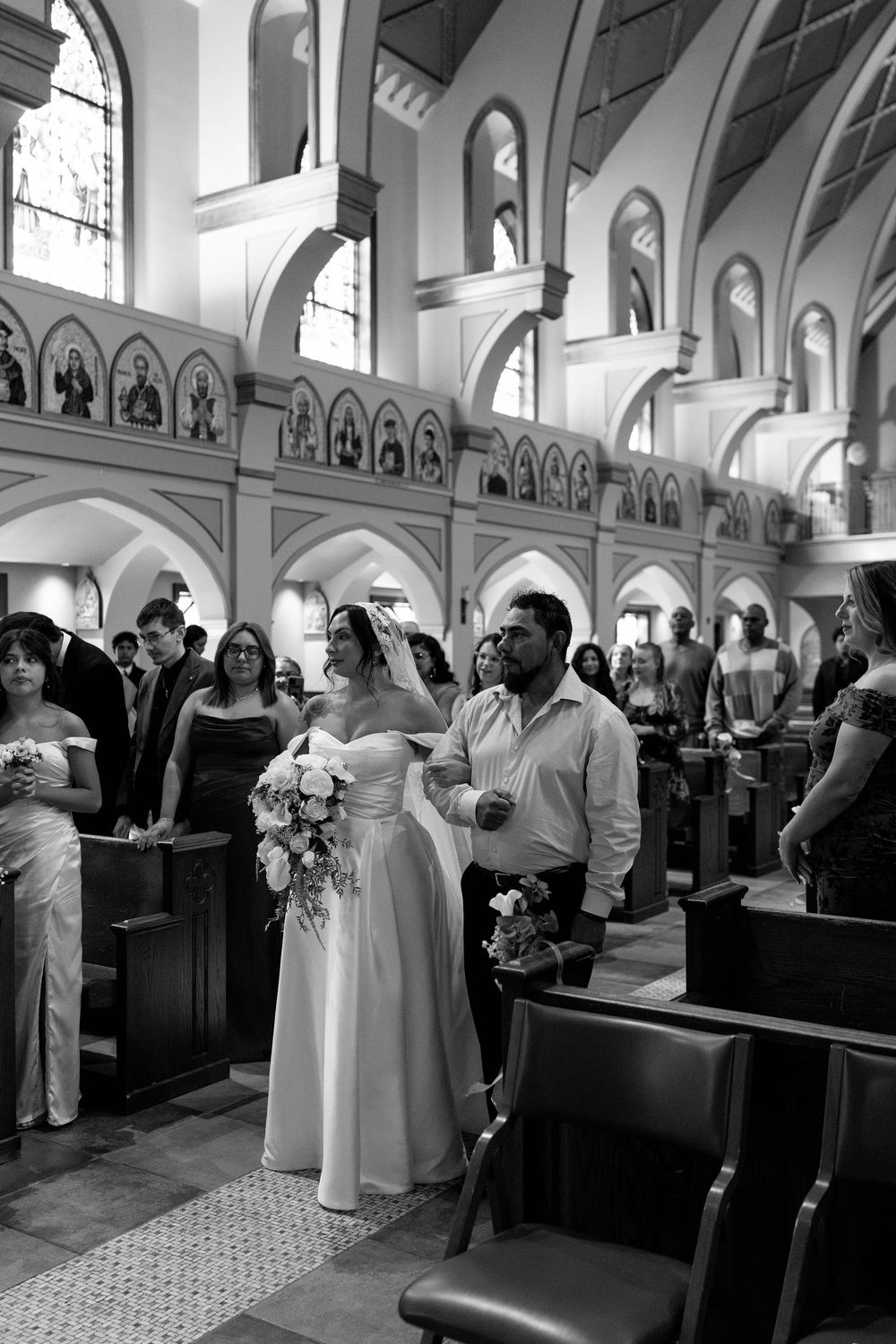 Bride in a white gown and veil walks down the church aisle on a man's arm as wedding guests look on.