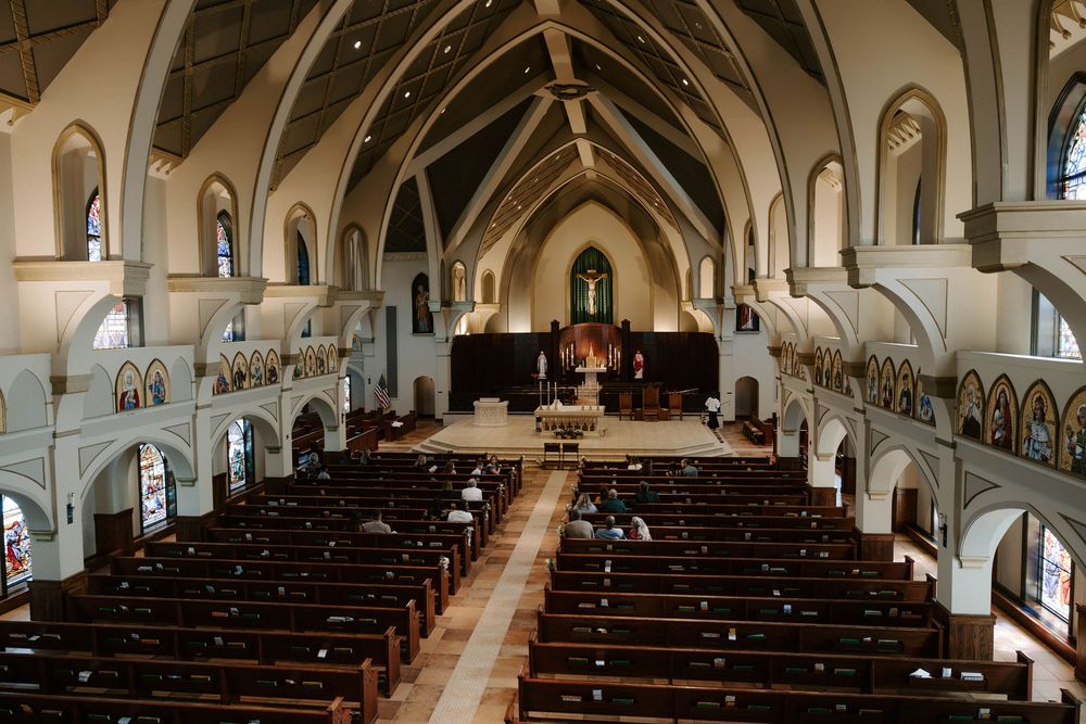 A wide view of a church interior with long wooden pews, vaulted ceiling, and a distant altar framed by stained glass windows.