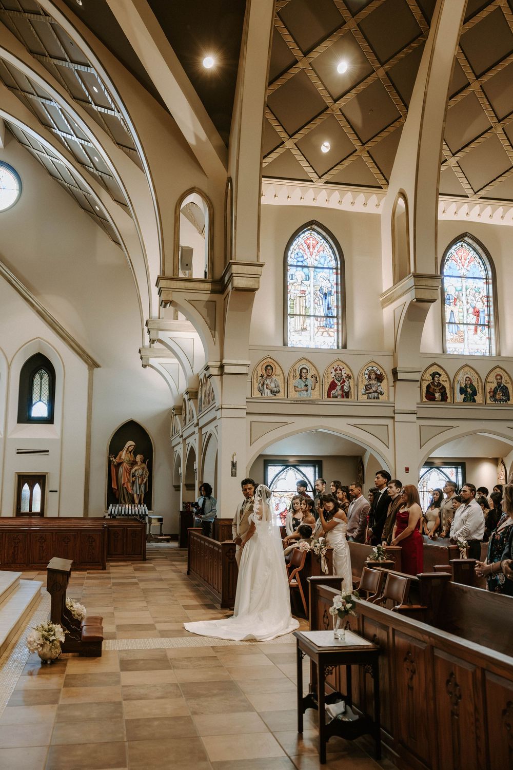 Bride and groom stand at the front of a church as wedding guests fill the pews beneath stained glass windows.