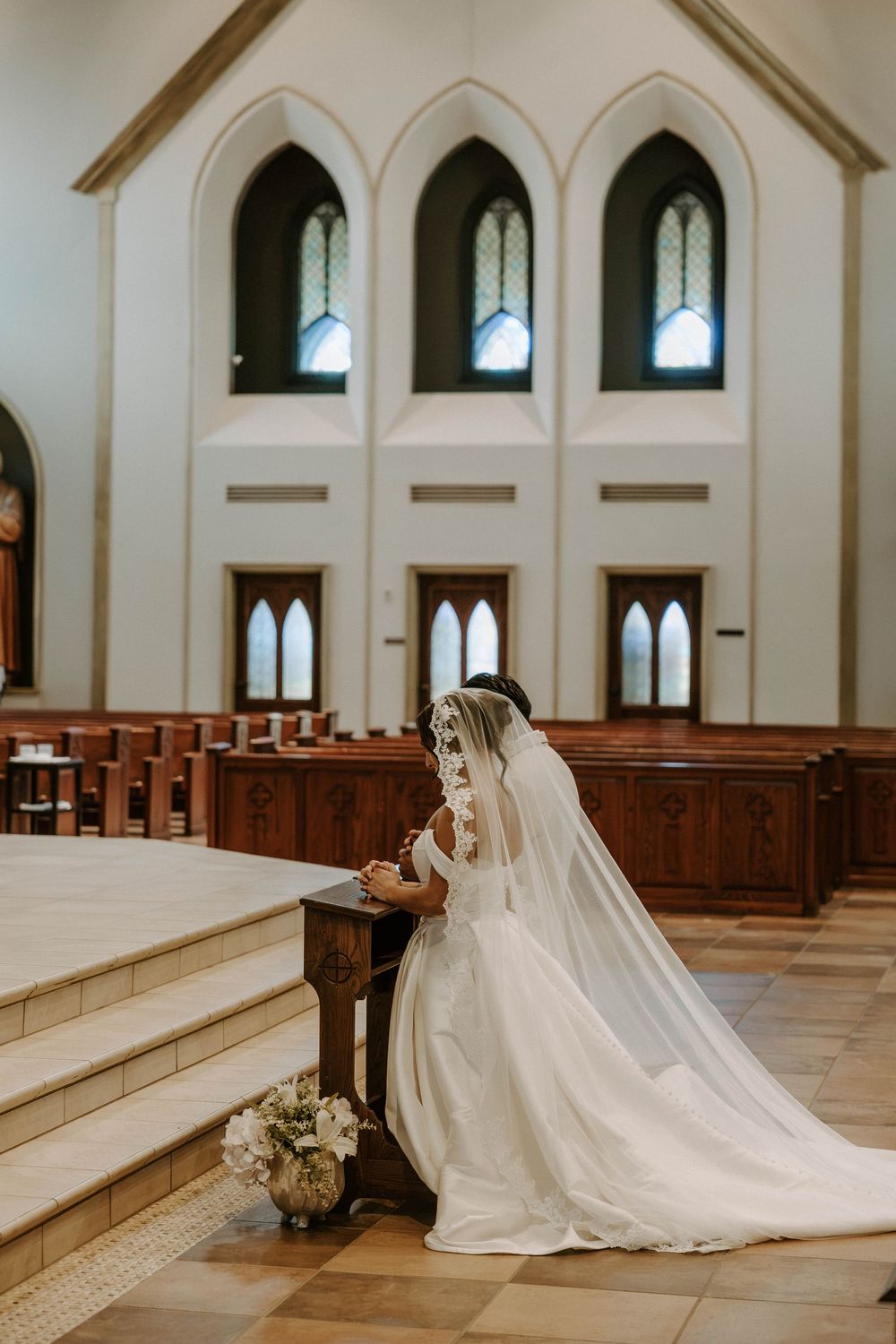 Bride in a white gown and long veil kneels in prayer at the front of a church near the altar steps.